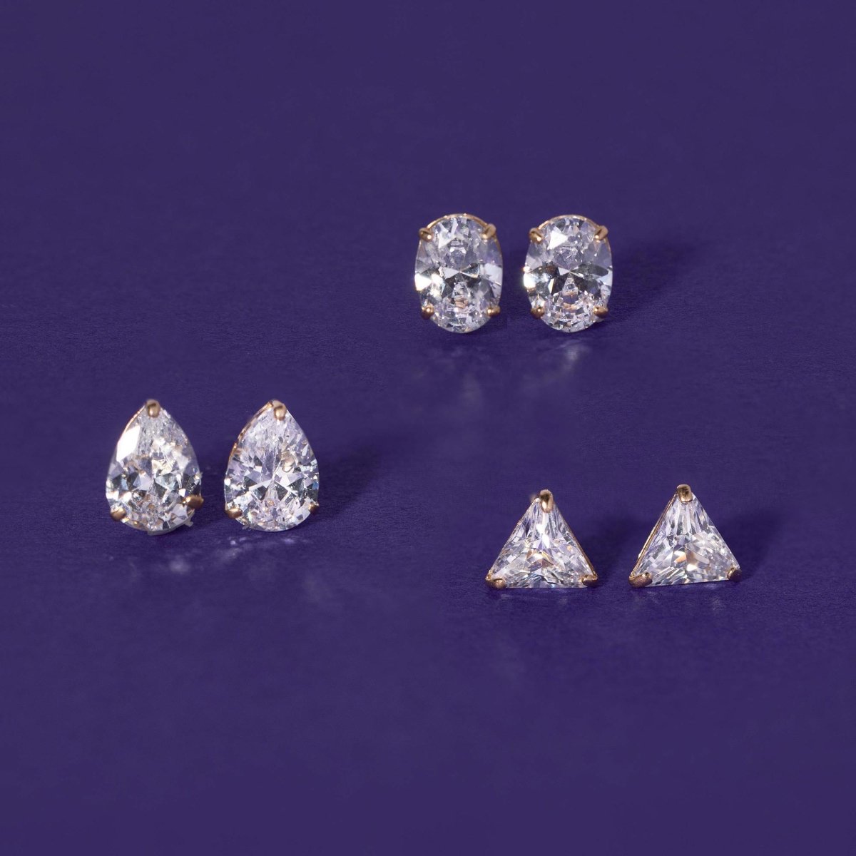 14K gold oval, pear and triangle studs with clear white zirconia stones, on a purple background