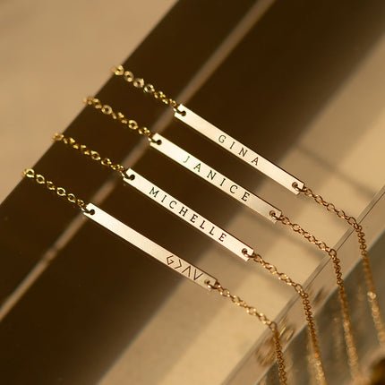 Four gold-filled thin bar bracelets engraved with names and a symbol, lined up on a reflective surface