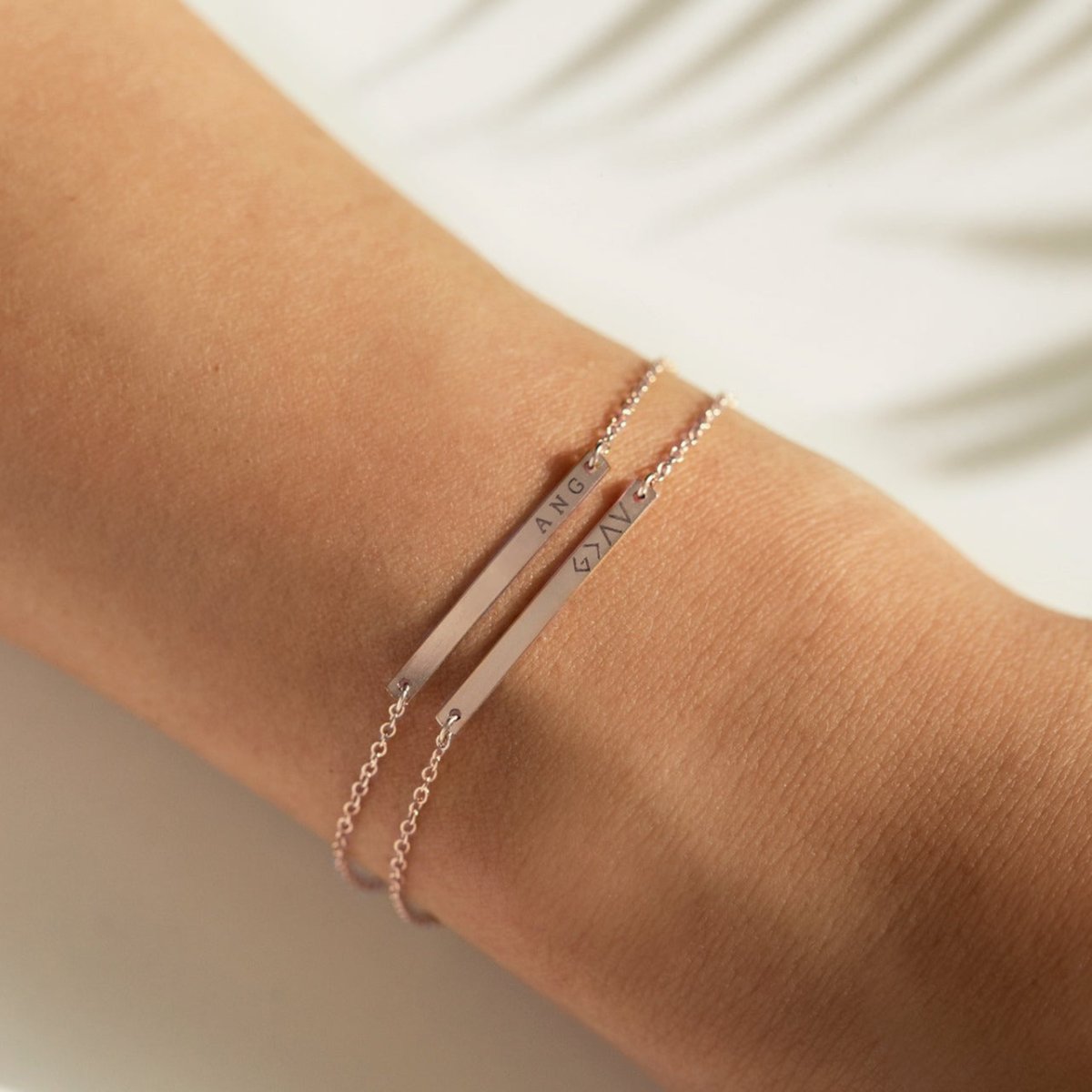 Two sterling silver thin bar bracelets engraved with a name and a symbol, shown layered on a wrist