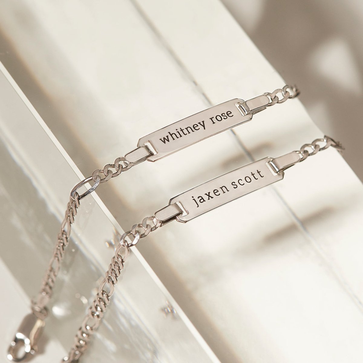 Sterling silver Figaro chain ID bracelets engraved with names, laid on a white wooden surface