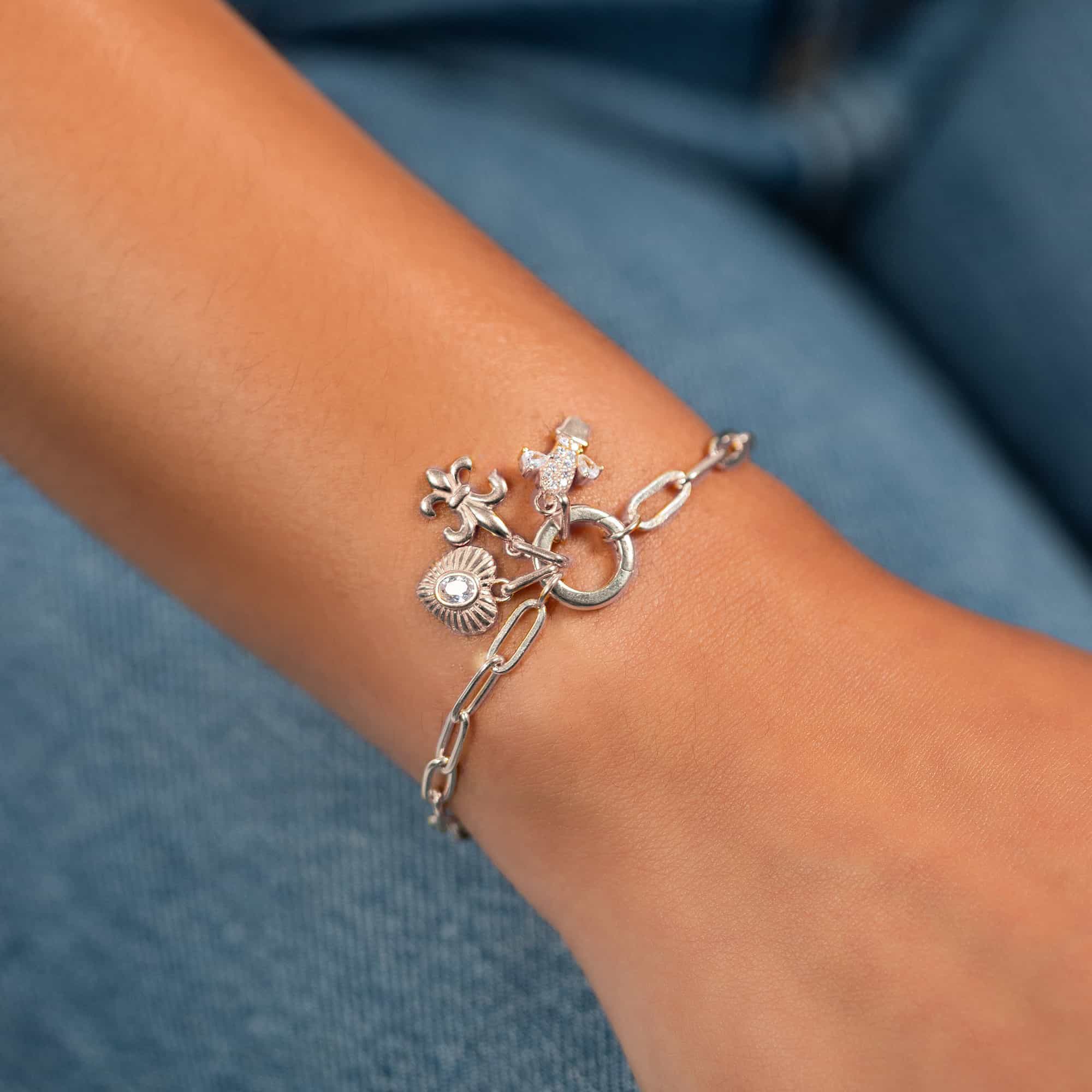 Silver paperclip charm bracelet with clear-stone, fleur-de-lis and heart charms on a wrist