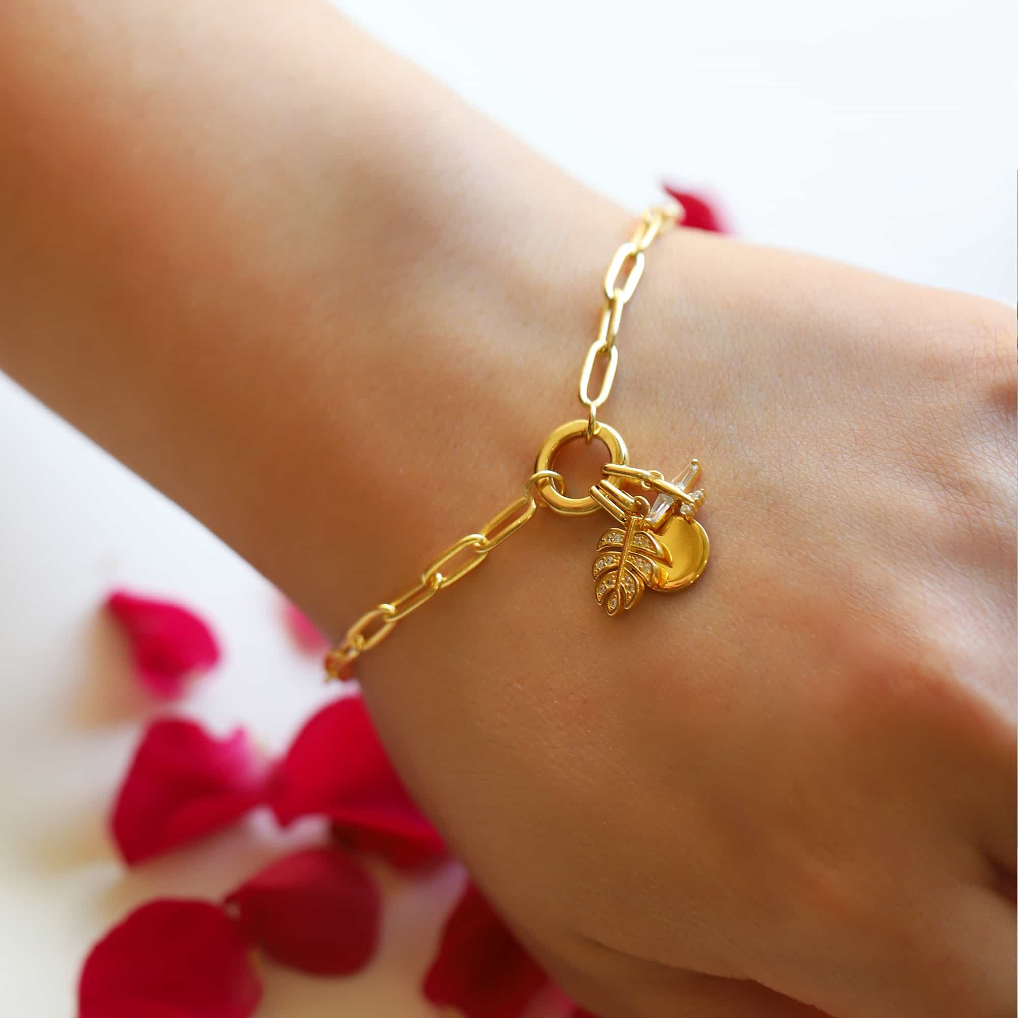 Gold paperclip chain charm bracelet with a pavé leaf and disc charms, shown on a wrist with rose petals