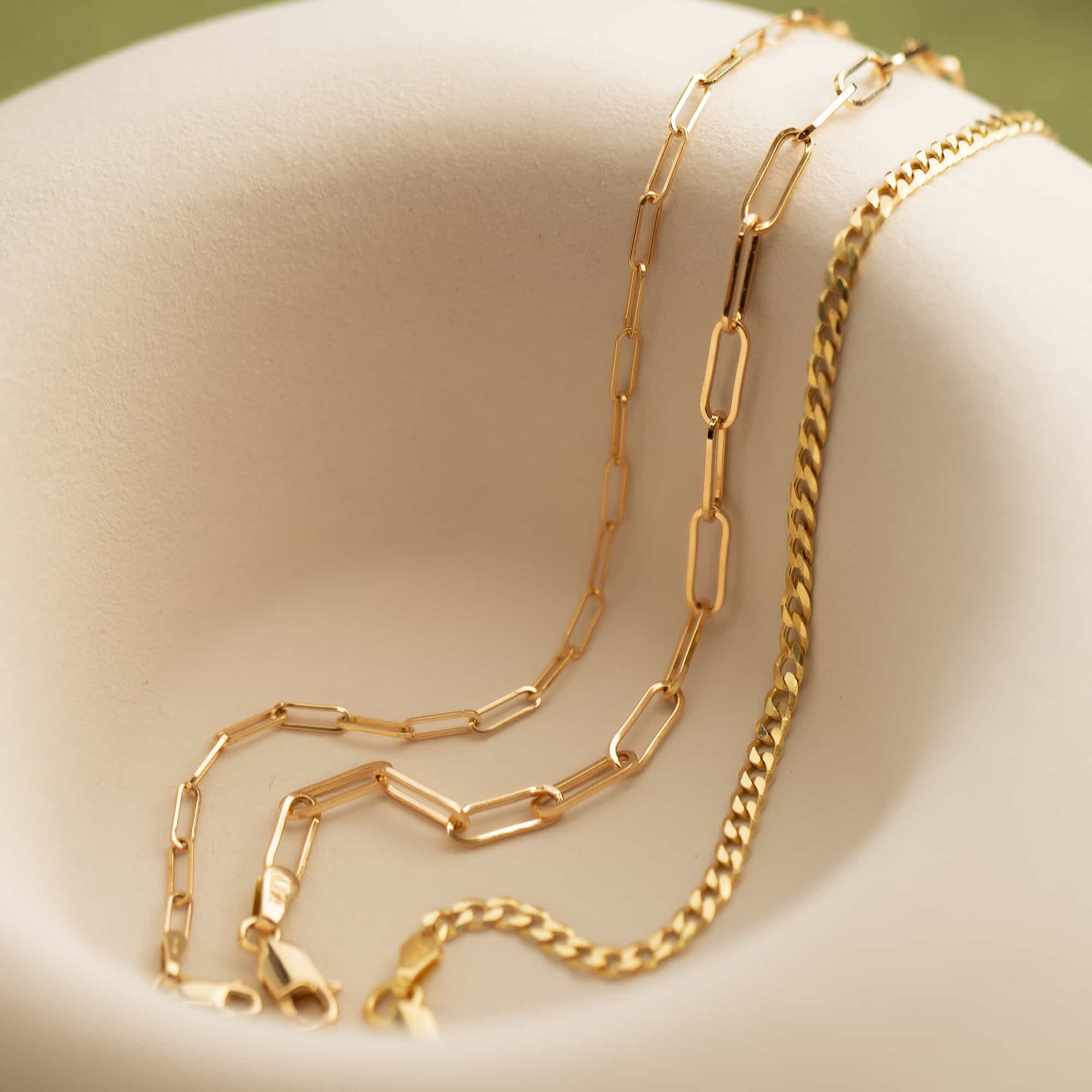 Gold paperclip chain and Cuban curb chain bracelets with lobster clasps, on a cream dish