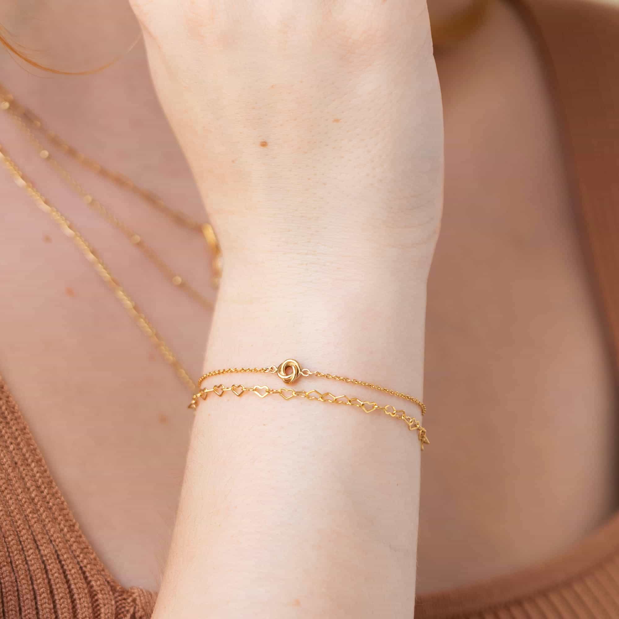 Gold-filled love knot chain bracelet worn layered on a model's wrist with a heart-link chain