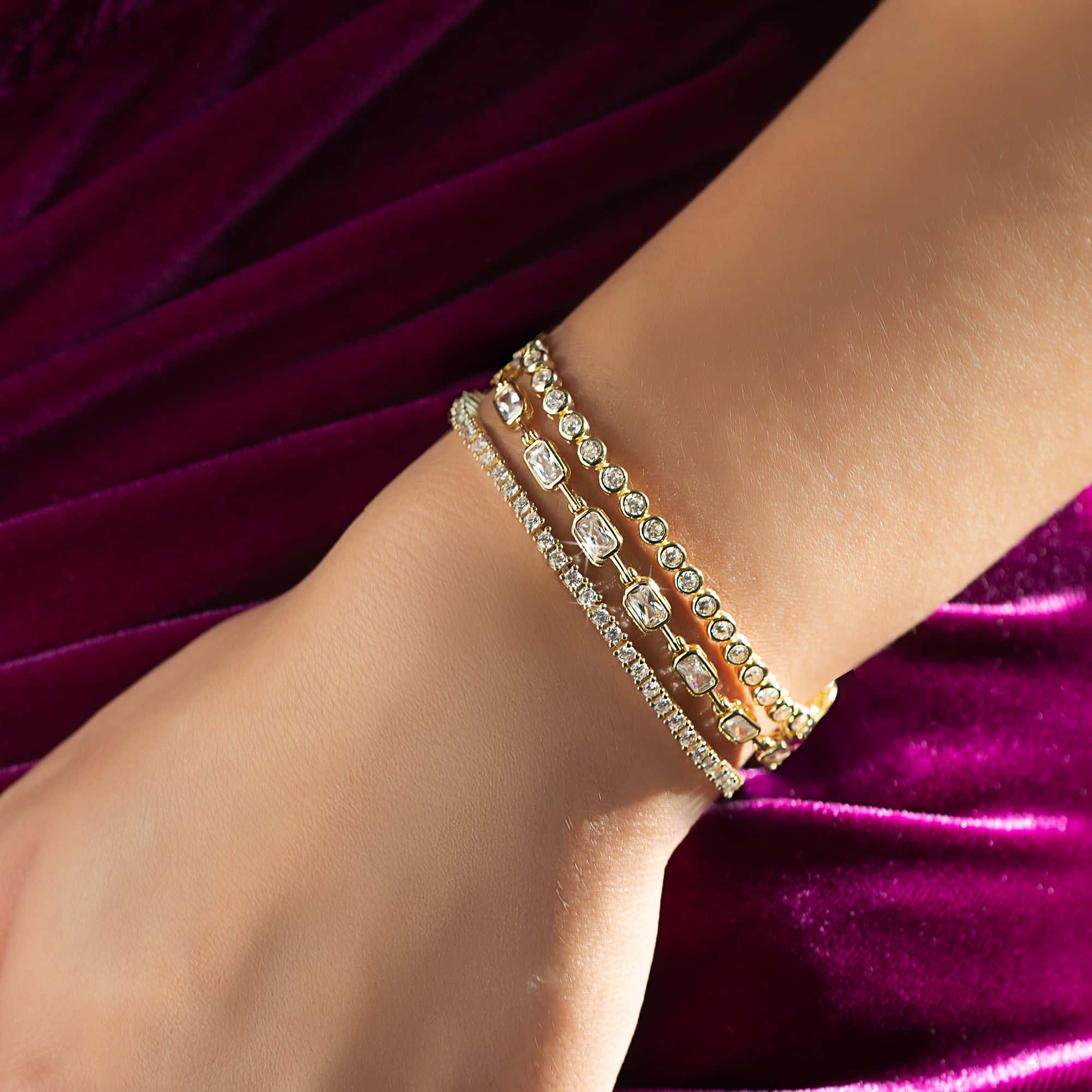 Gold clear-stone tennis bracelets stacked on a model's wrist against magenta velvet
