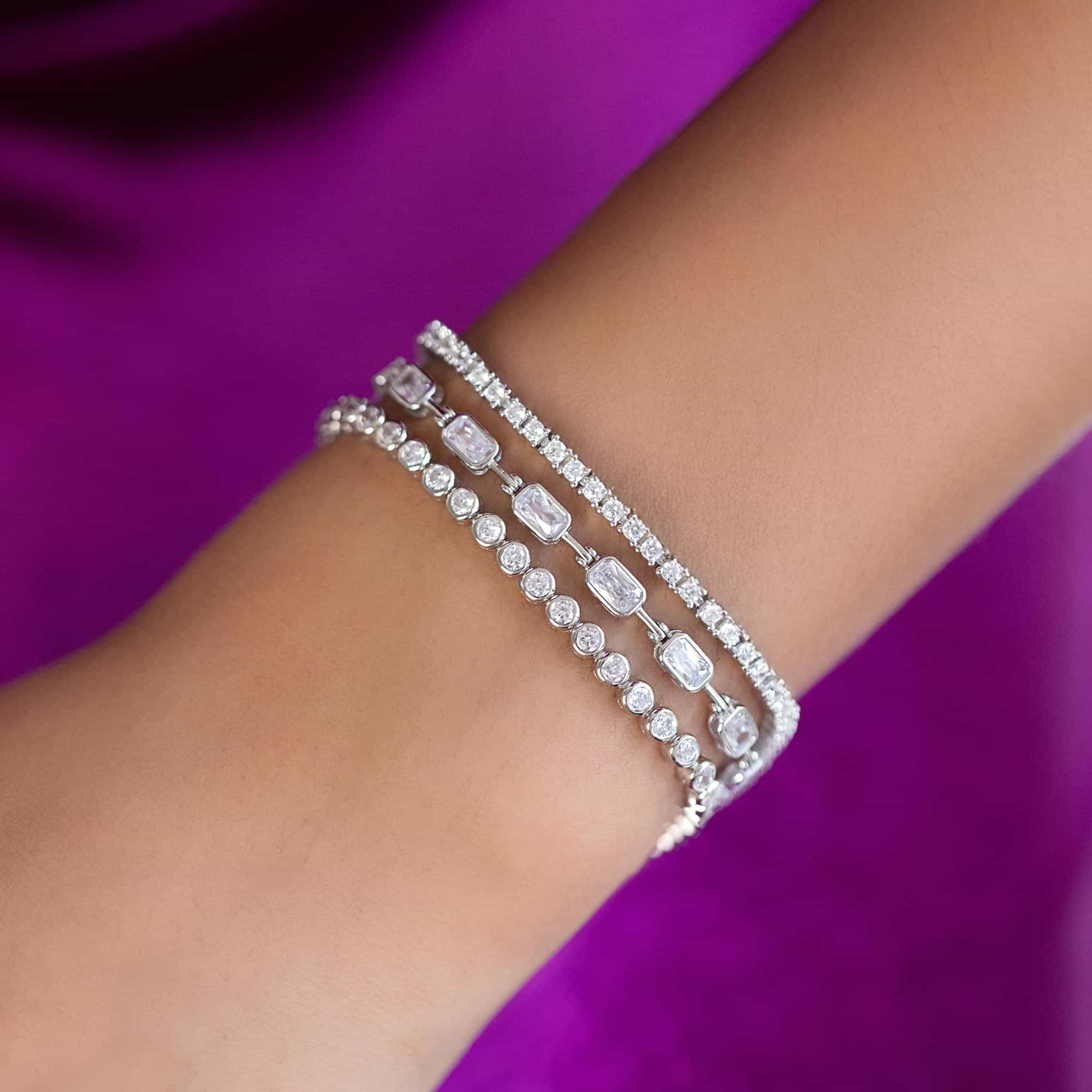 Silver clear-stone tennis bracelets in a stack on a model's wrist against purple