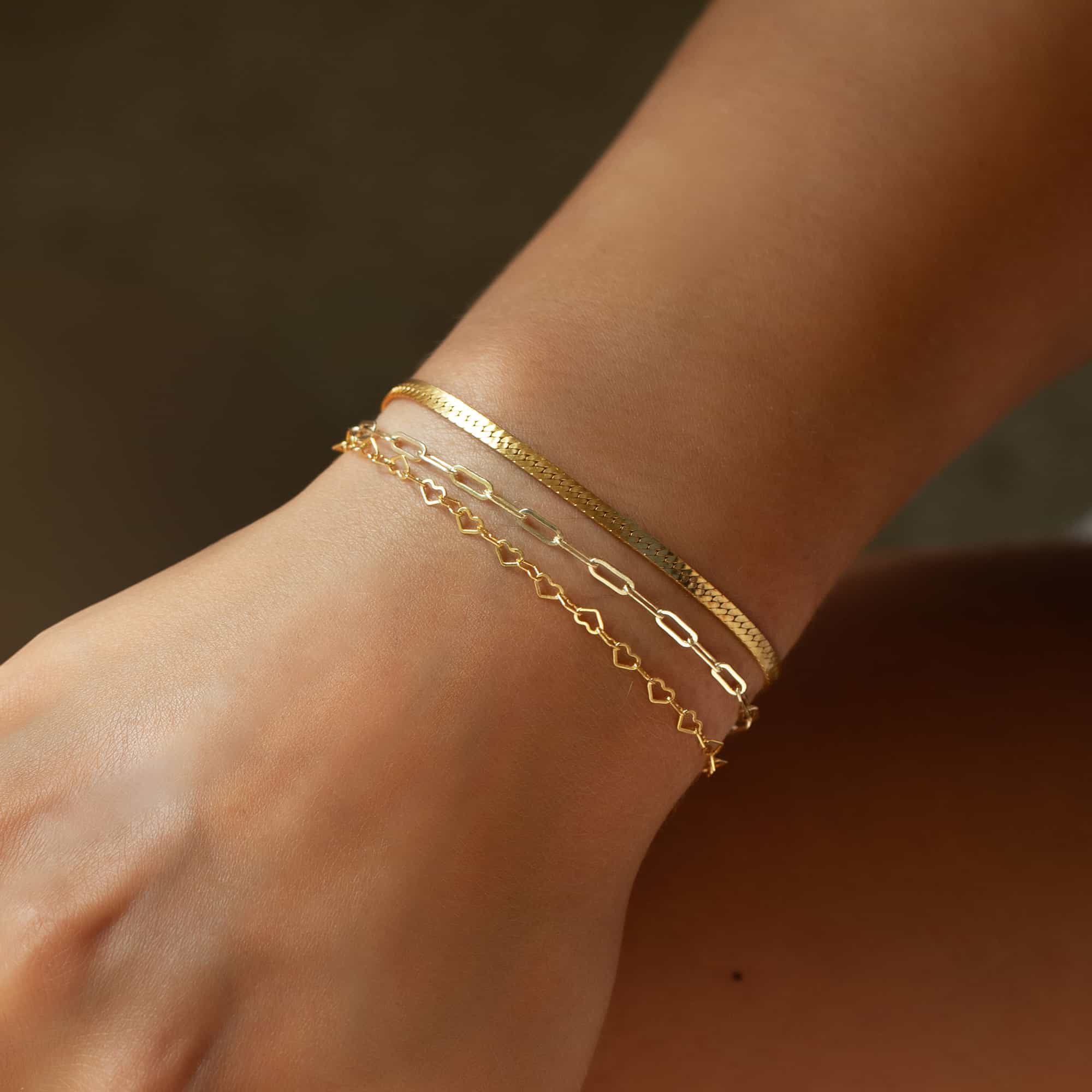 Gold-filled paperclip chain bracelet layered with a herringbone and heart-link chain on a wrist