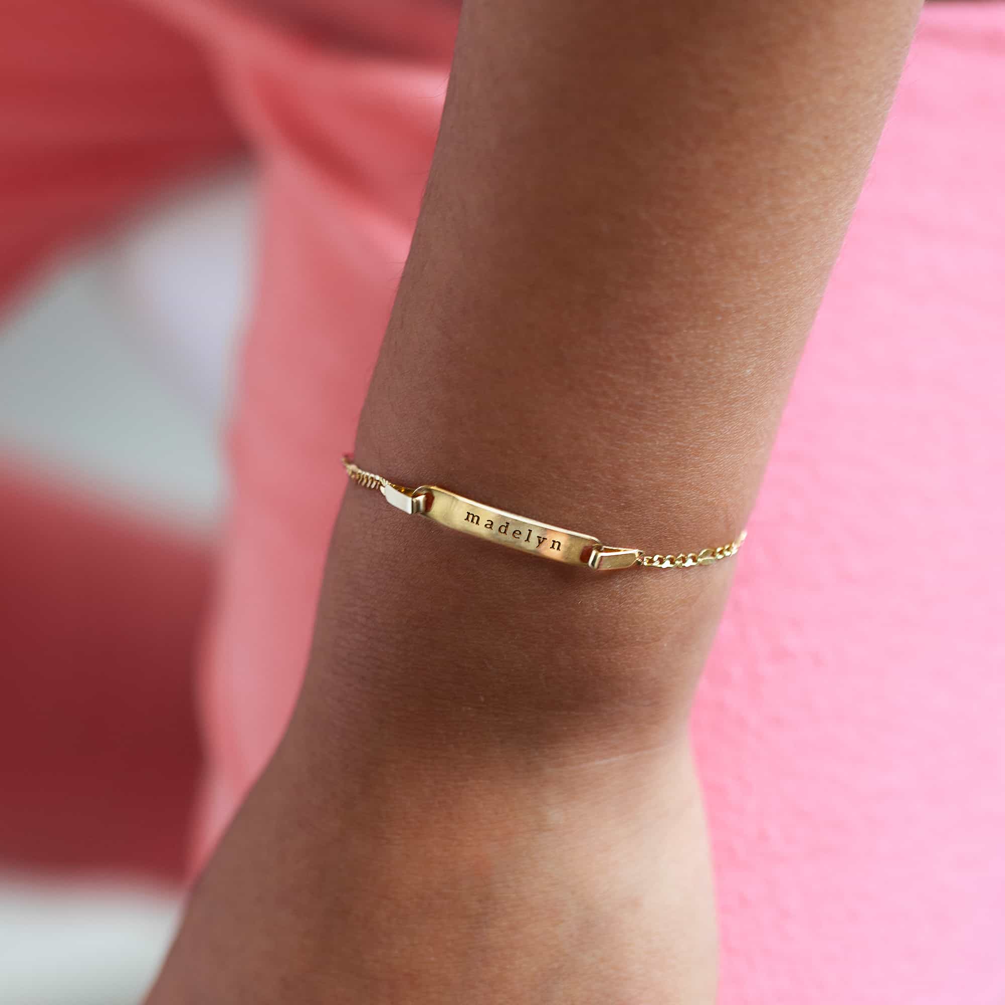 14K gold ID bracelet engraved with a name, shown on a child's wrist against pink