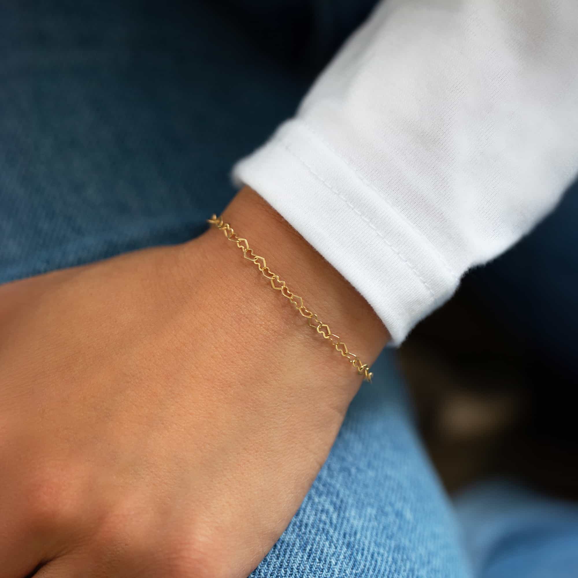 14K gold delicate open heart-link chain bracelet, shown on a model's wrist over denim