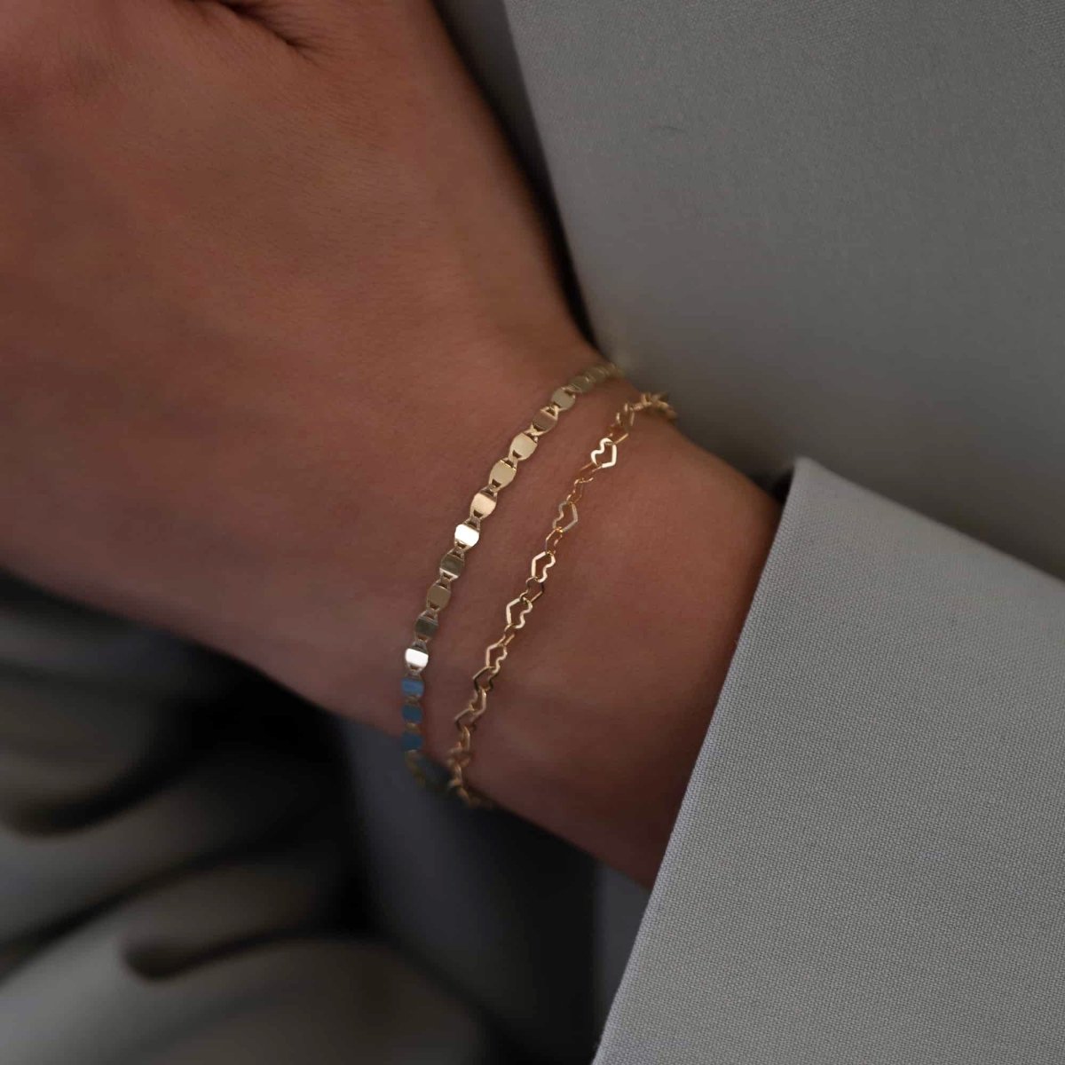 14K gold heart-link chain bracelet layered with a flat disc chain, shown on a model's wrist