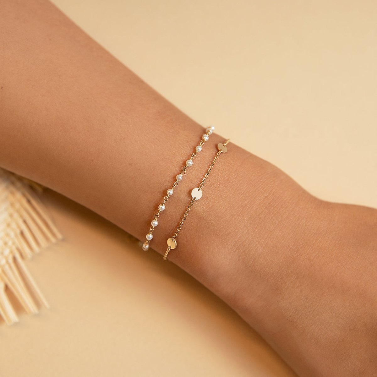 14K gold eternity disc bracelet stacked with a pearl bracelet on a wrist, on a cream background