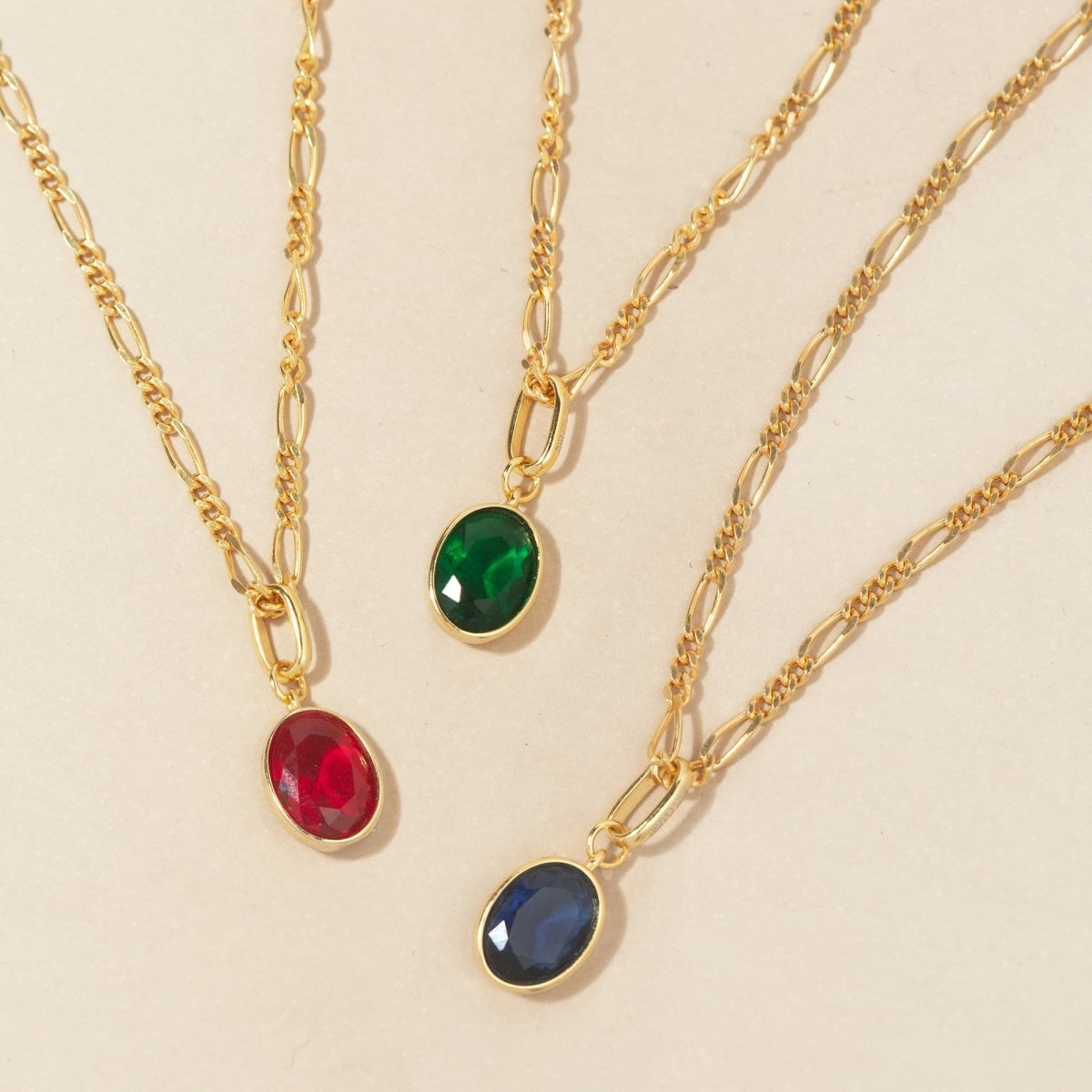 Gold vermeil oval green emerald charm on a Figaro chain beside red and blue charms, on a cream background