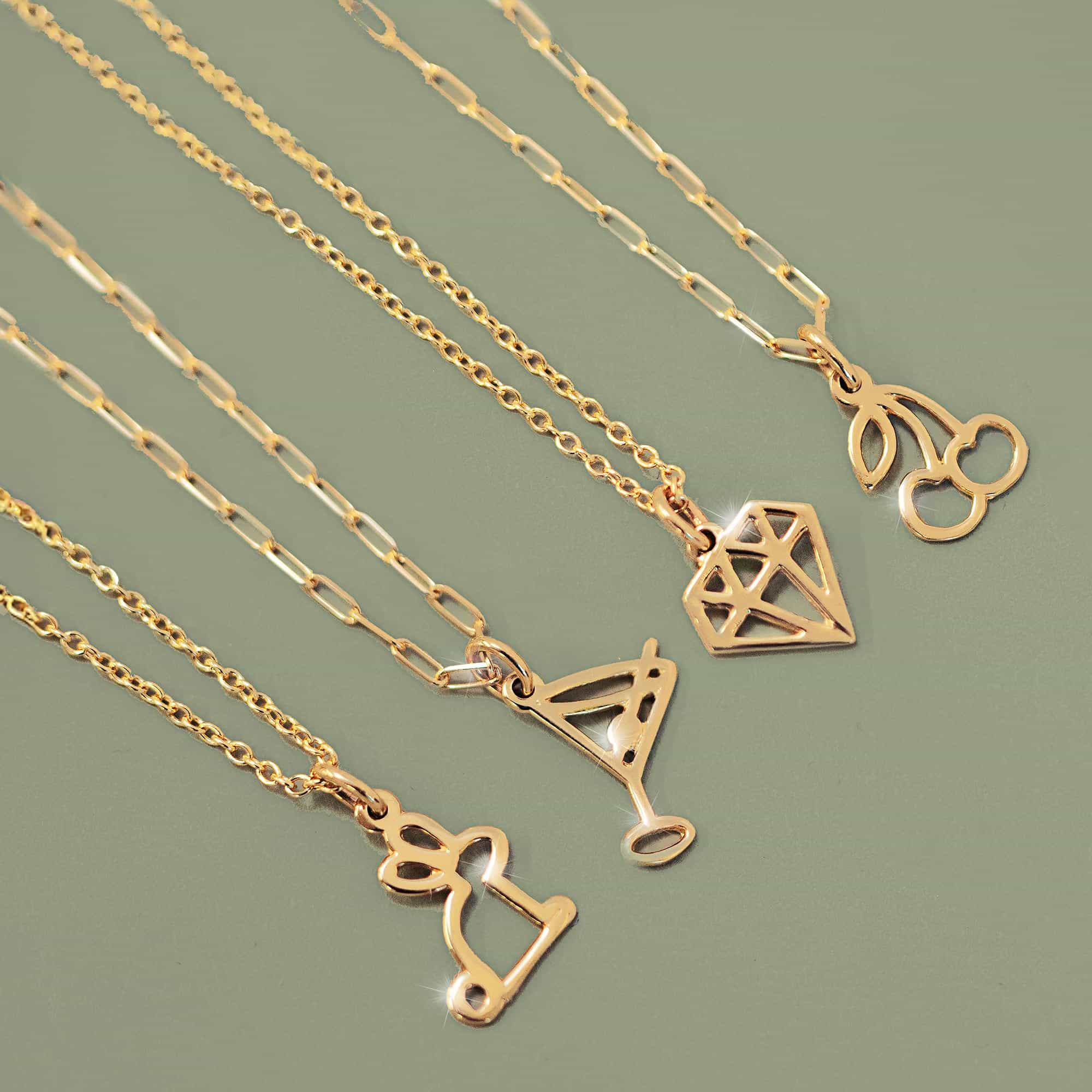 Four gold charm necklaces with cherry, martini glass, diamond outline and cocktail charms, on a sage background