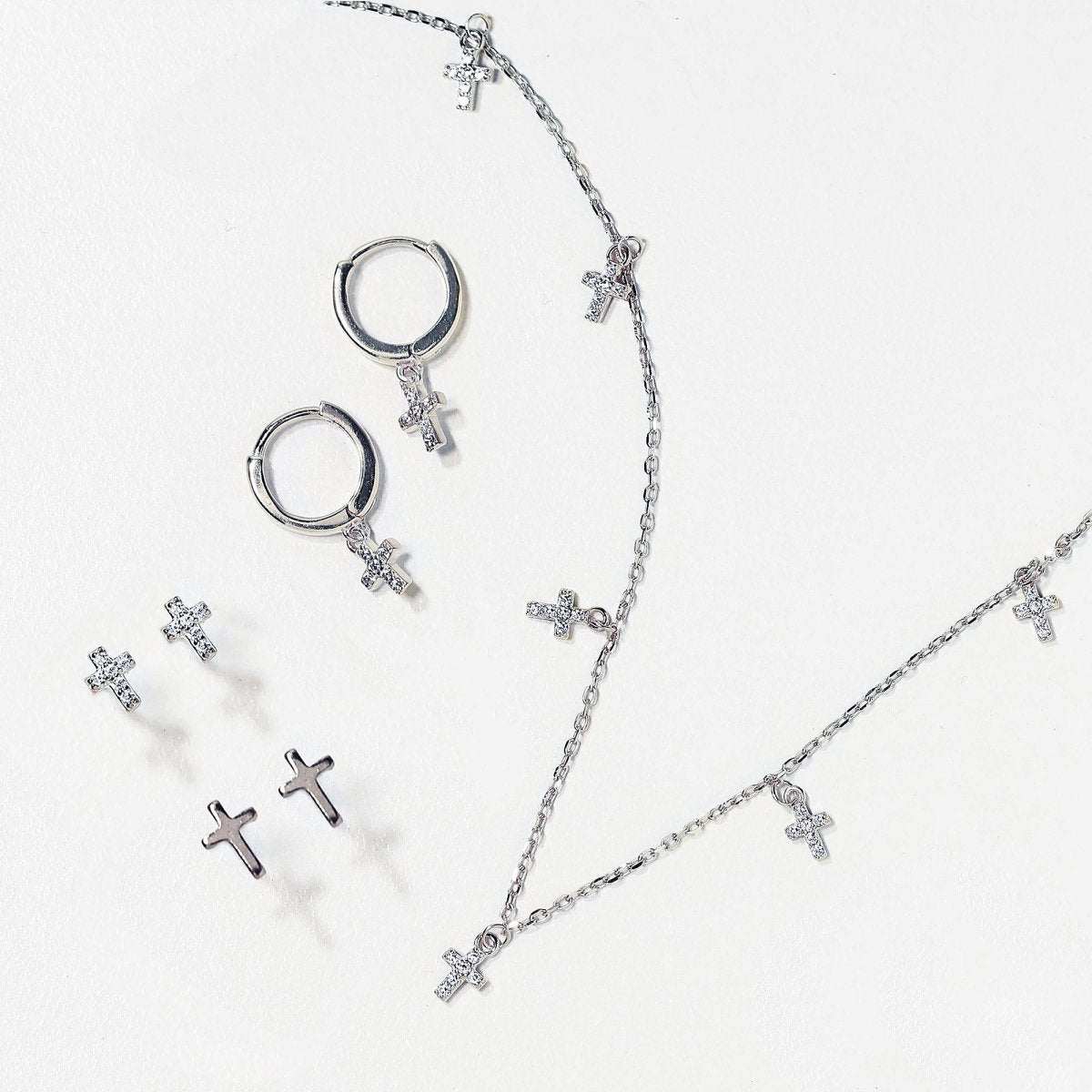 Silver cross studs, huggie hoops and chains laid out together on a white background