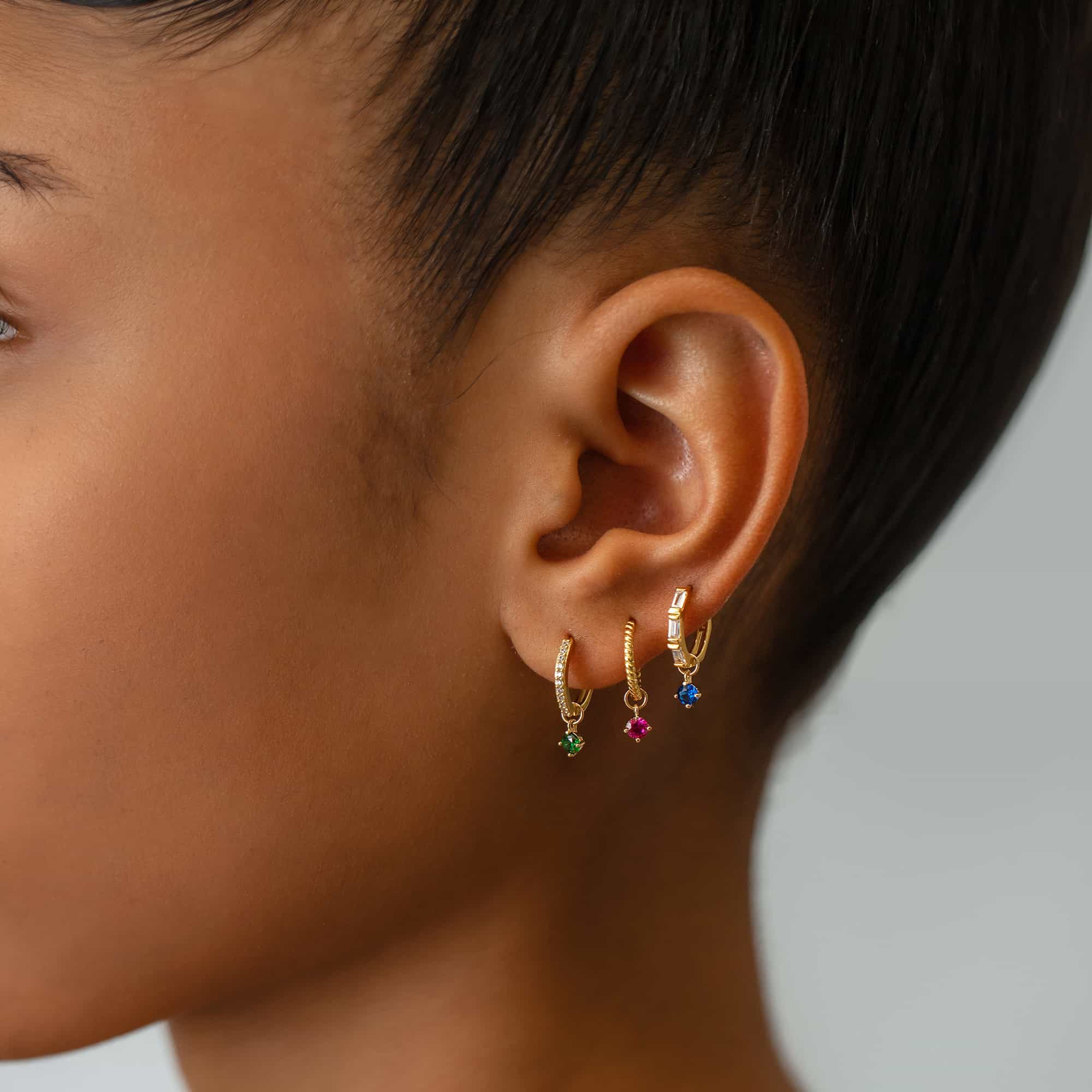 14K gold huggie hoops with dangling colored stone charms, shown on a model's ear