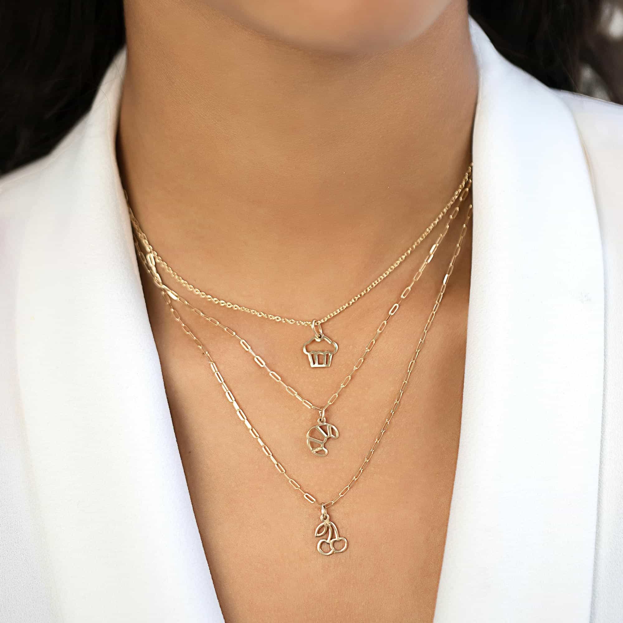 Three layered gold chains with cupcake, croissant and cherry charm pendants, shown on a model's neckline