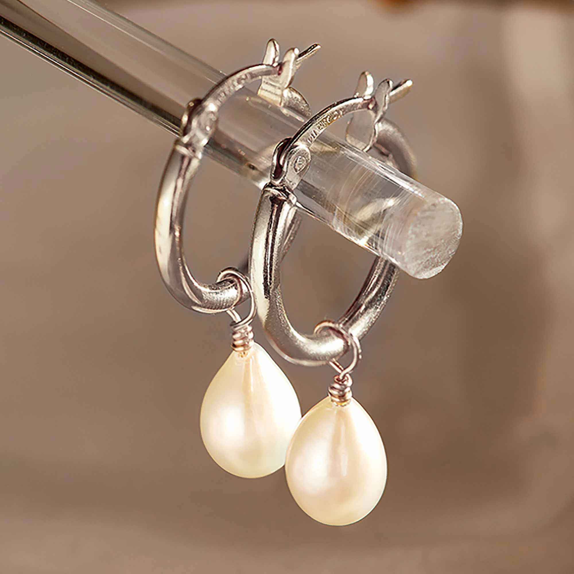 Sterling silver huggie hoops with a dangling white teardrop pearl, held on a clear rod, close-up