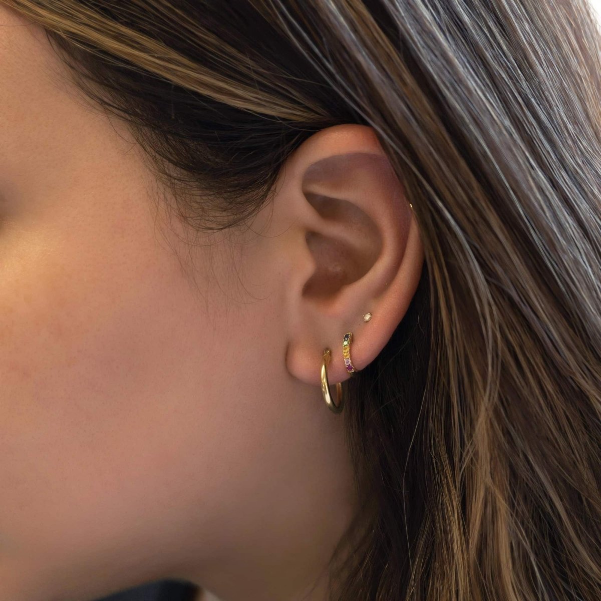 14K gold rainbow pavé huggie hoop shown on a model's lobe with a plain gold hoop