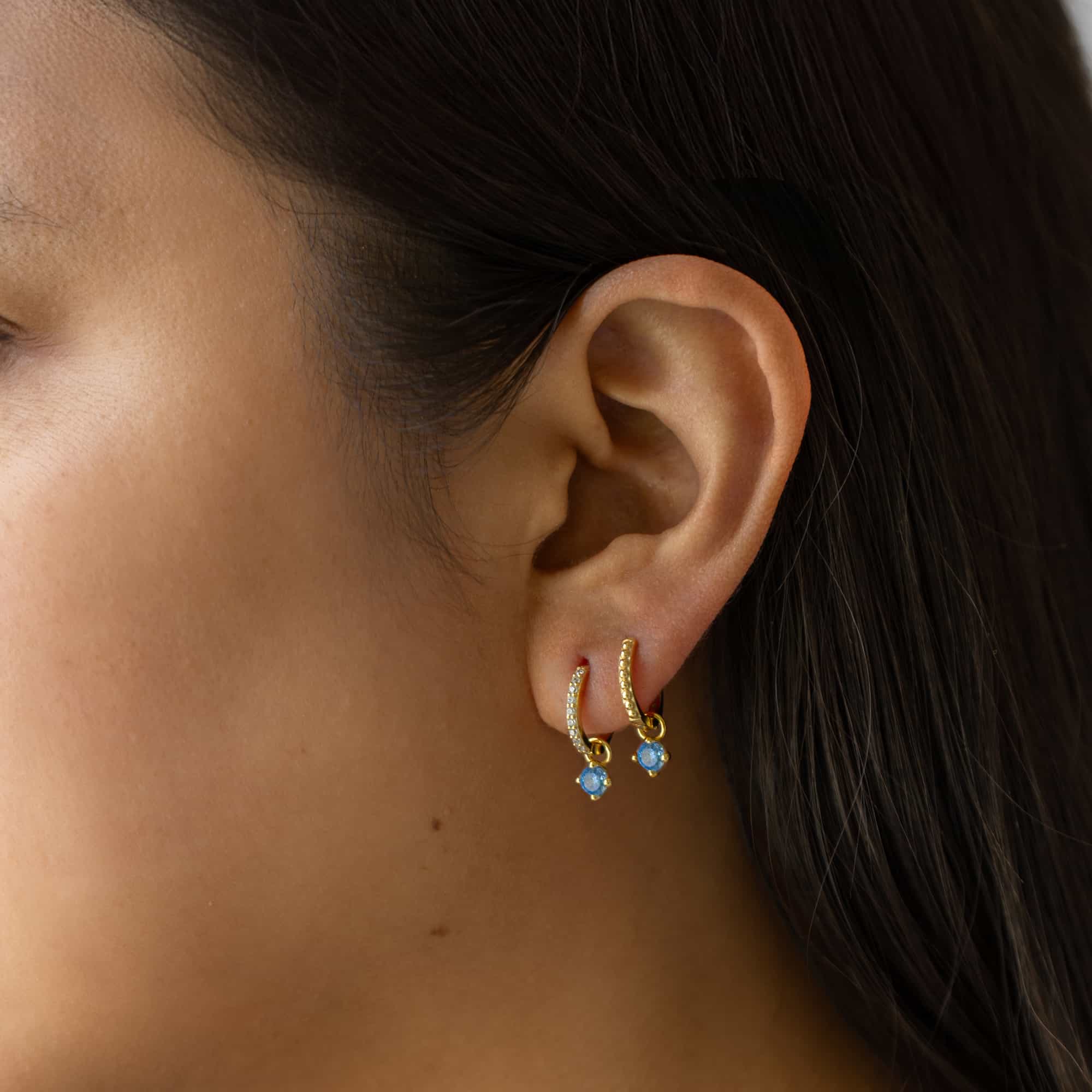 Gold vermeil pavé huggie hoops with dangling blue zircon charms, shown on a model's ear