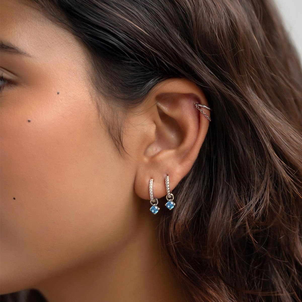 Sterling silver pavé huggie hoops with dangling blue zircon charms, shown on a model's stacked ear