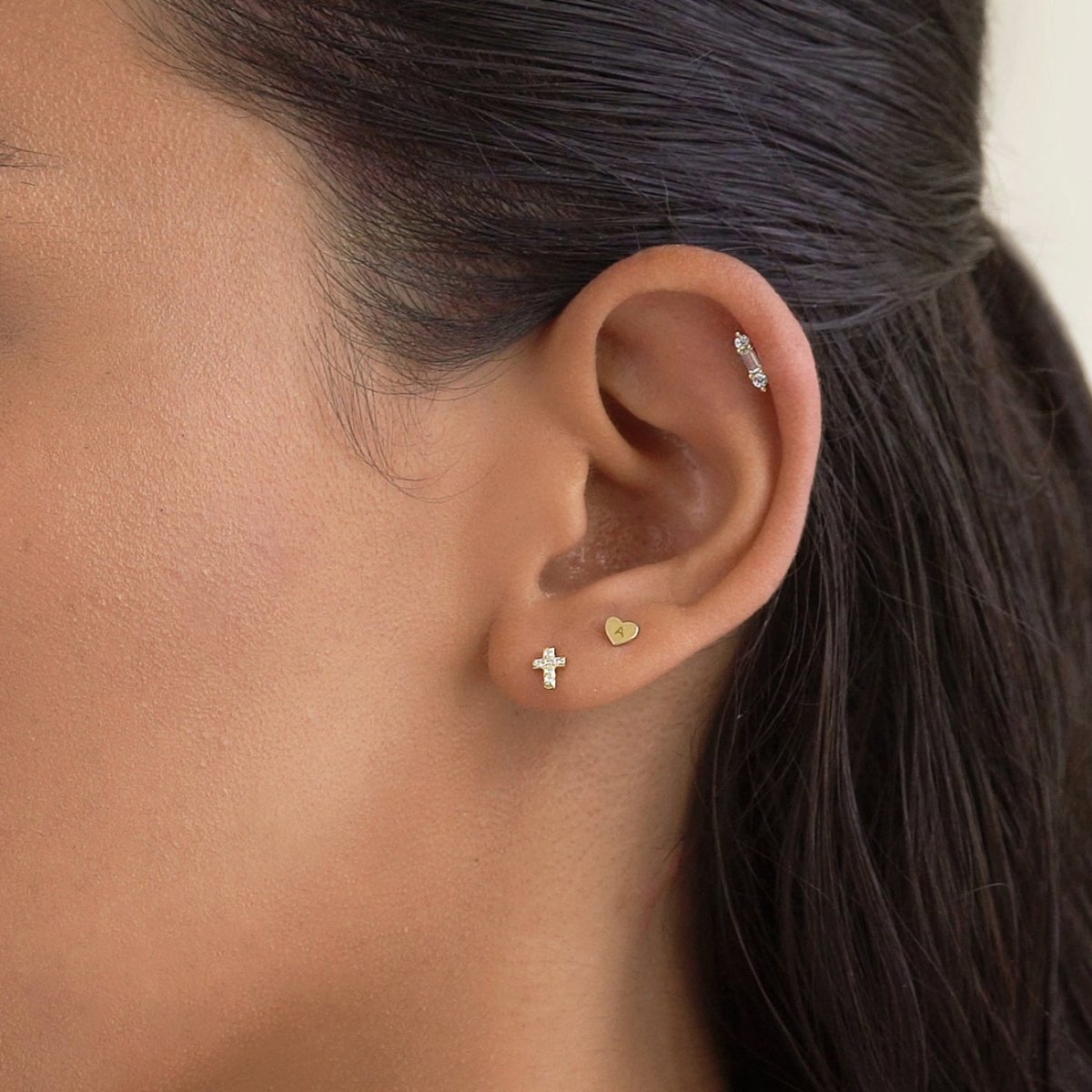 14K gold tiny heart initial stud shown on a model's earlobe beside a cross stud