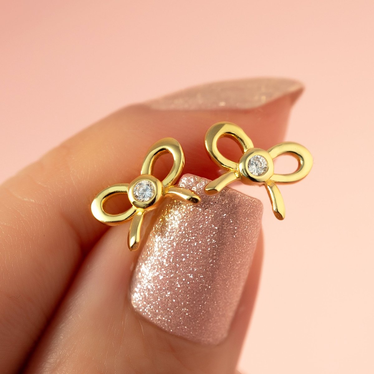 Gold vermeil bow studs with clear bezel stones held against a fingertip, close-up detail