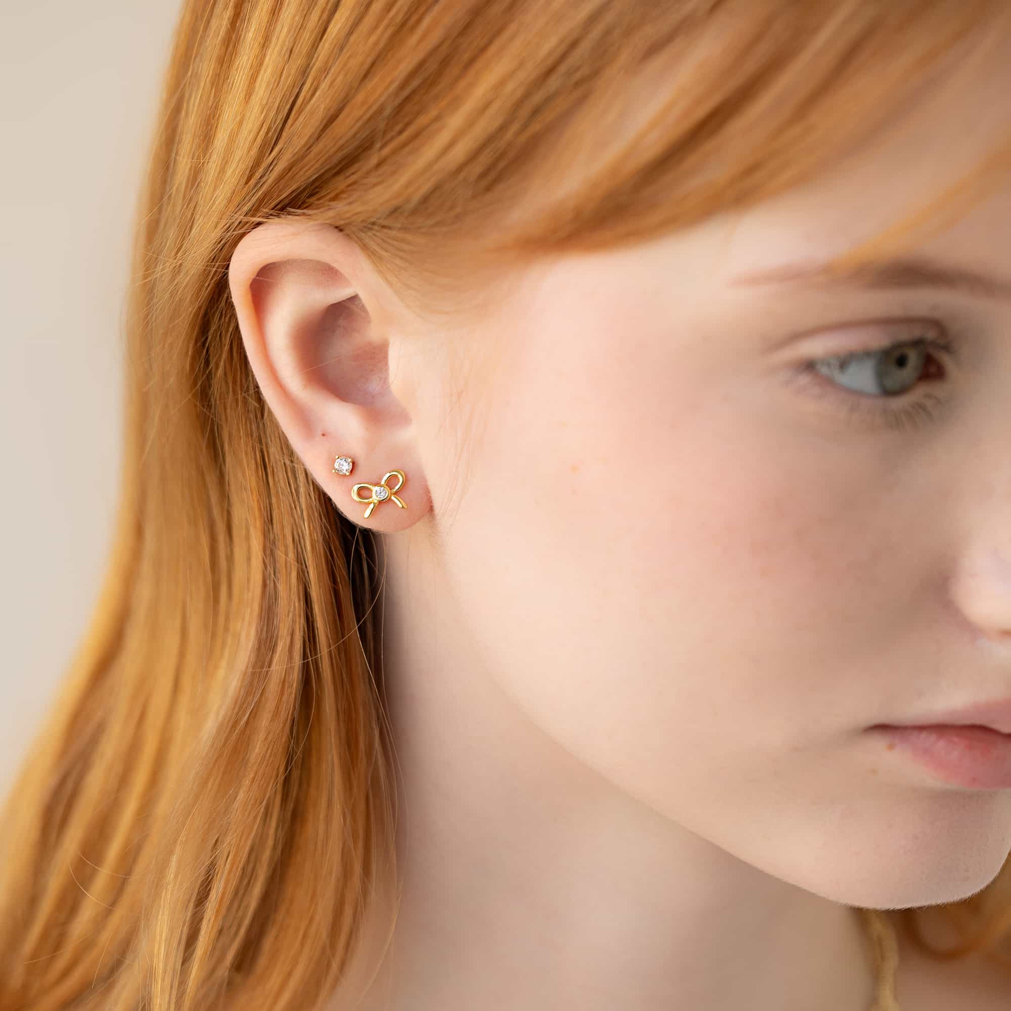 Gold vermeil crystal bow stud shown on a model's lobe beside a clear solitaire stud