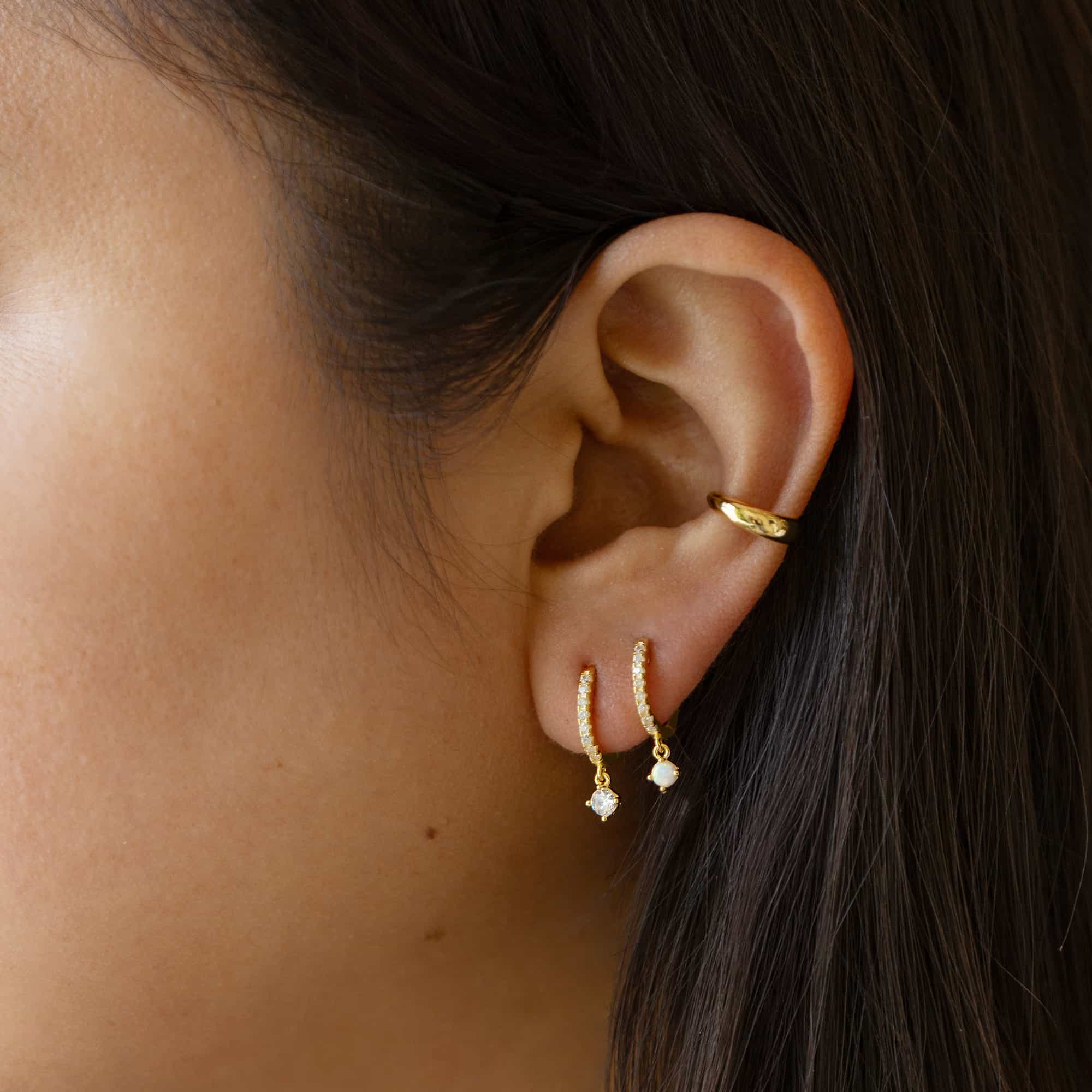 Gold vermeil pavé huggie hoops, one with a white opal drop and one with a clear stone drop, on a model's lobe