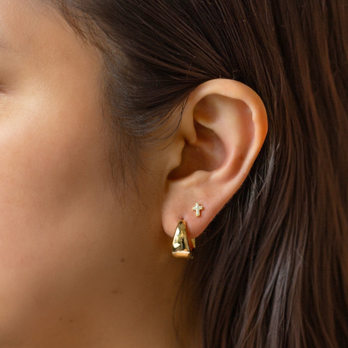 14K gold pavé zirconia cross stud shown on a model's lobe beside a gold huggie hoop