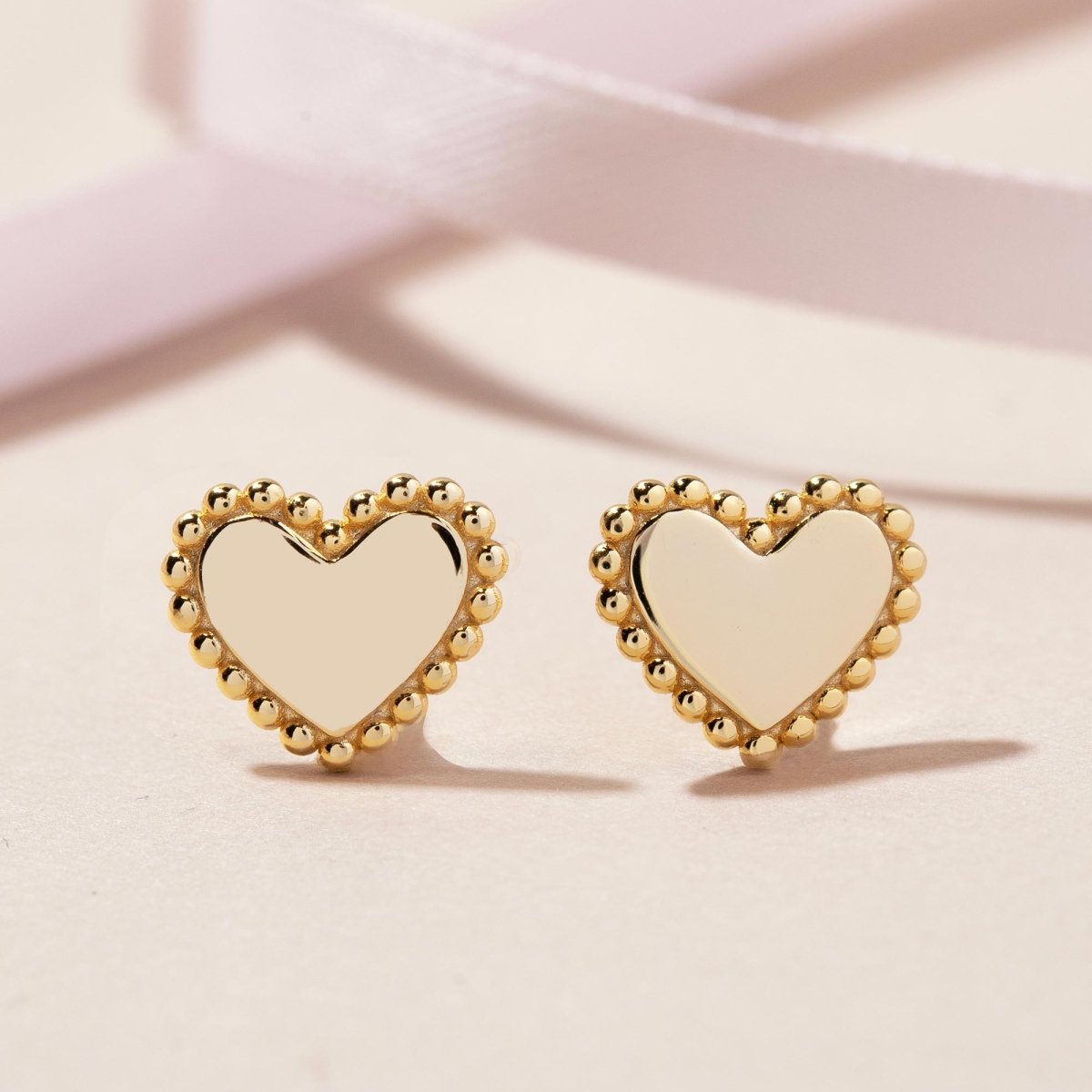 Gold vermeil heart stud earrings with beaded edges, on a pink ribbon background
