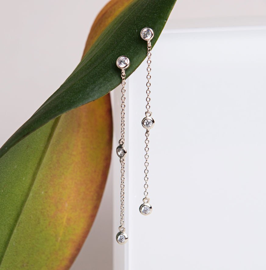 Sterling silver white zirconia drop earrings staggered along chain, resting against a green leaf