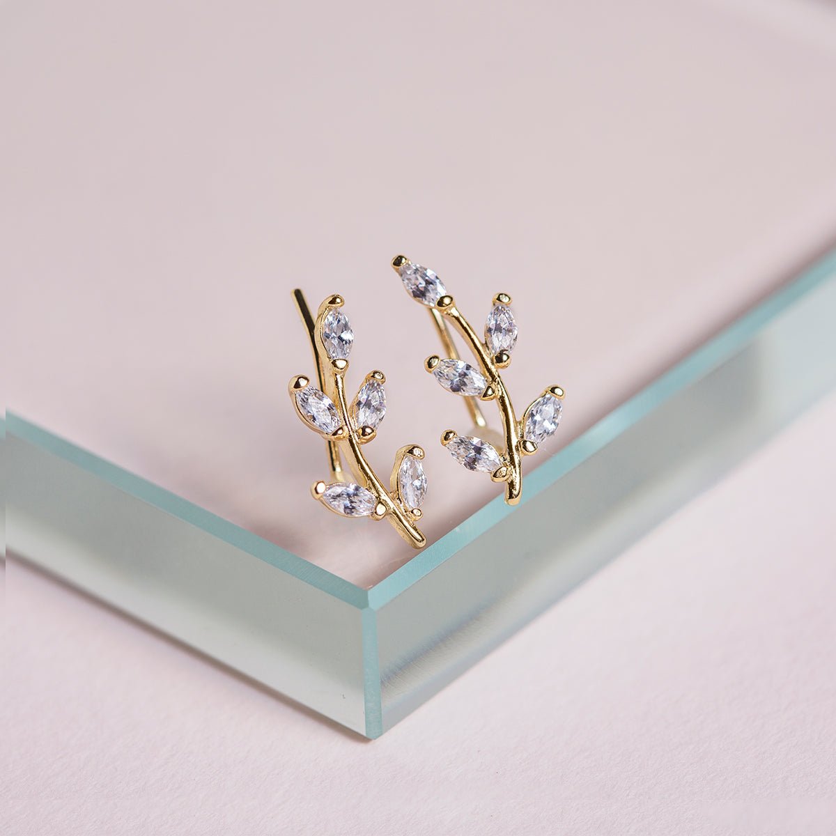 Gold vermeil leaf ear climbers with marquise clear stones, on a pale pink and glass surface