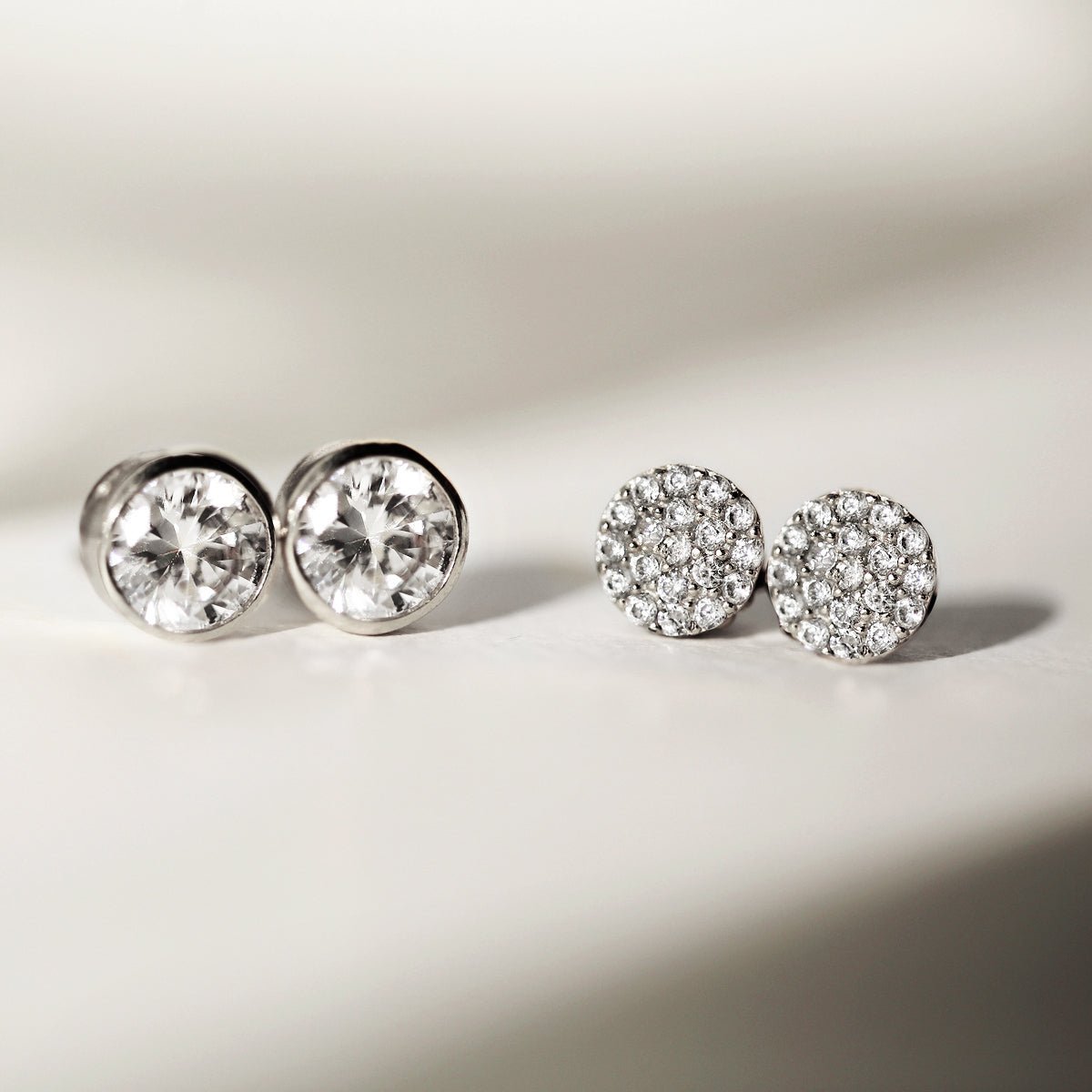 Sterling silver pavé round studs with white zirconia beside solitaire studs, on a cream background