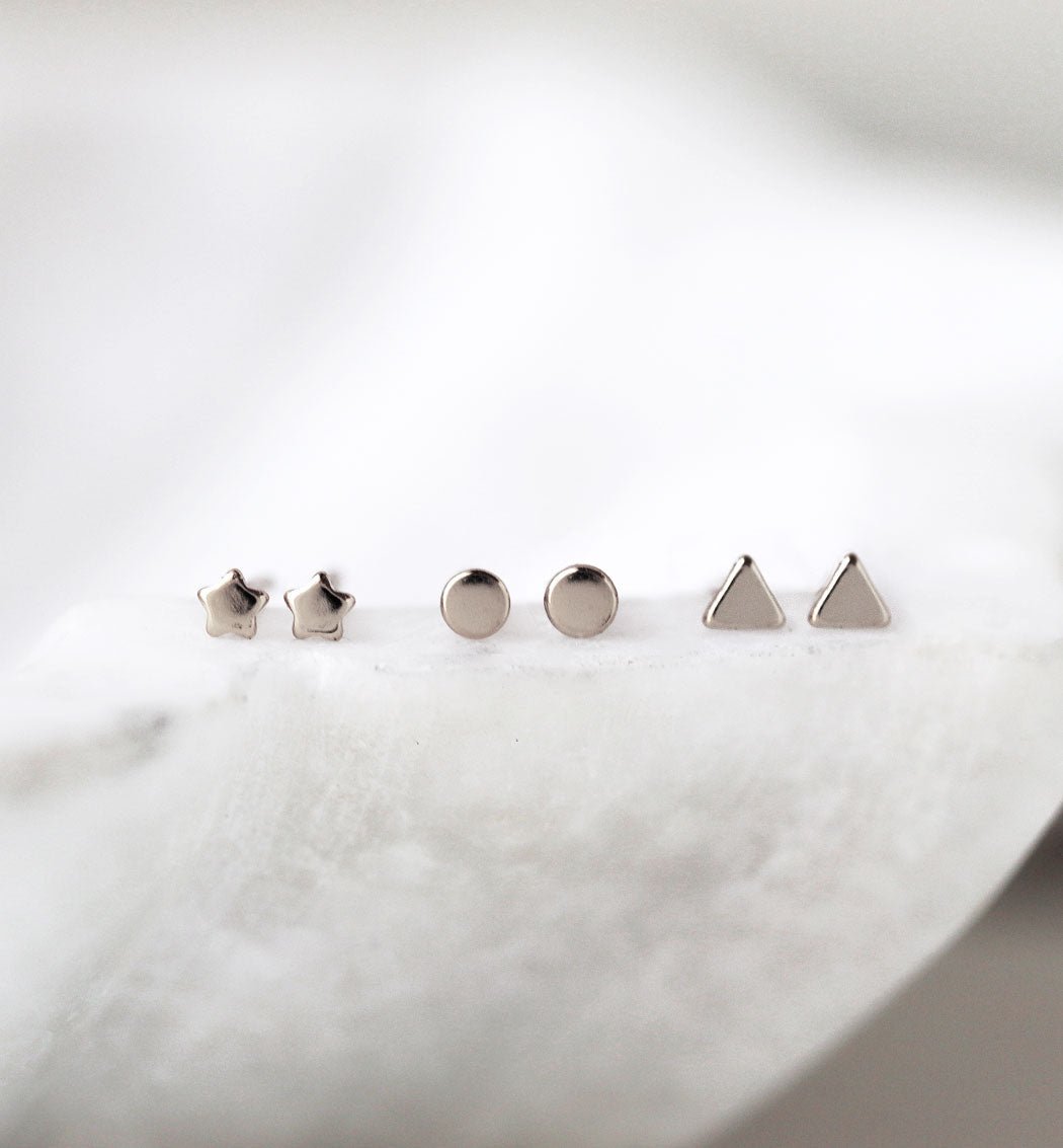 Sterling silver tiny star, circle and triangle studs lined up on white marble, close-up detail