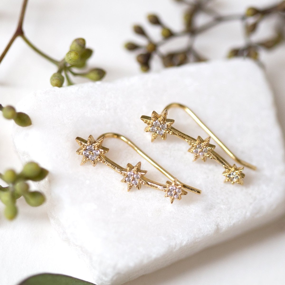 Gold vermeil star ear climbers with white zirconia, close-up detail on white marble with greenery