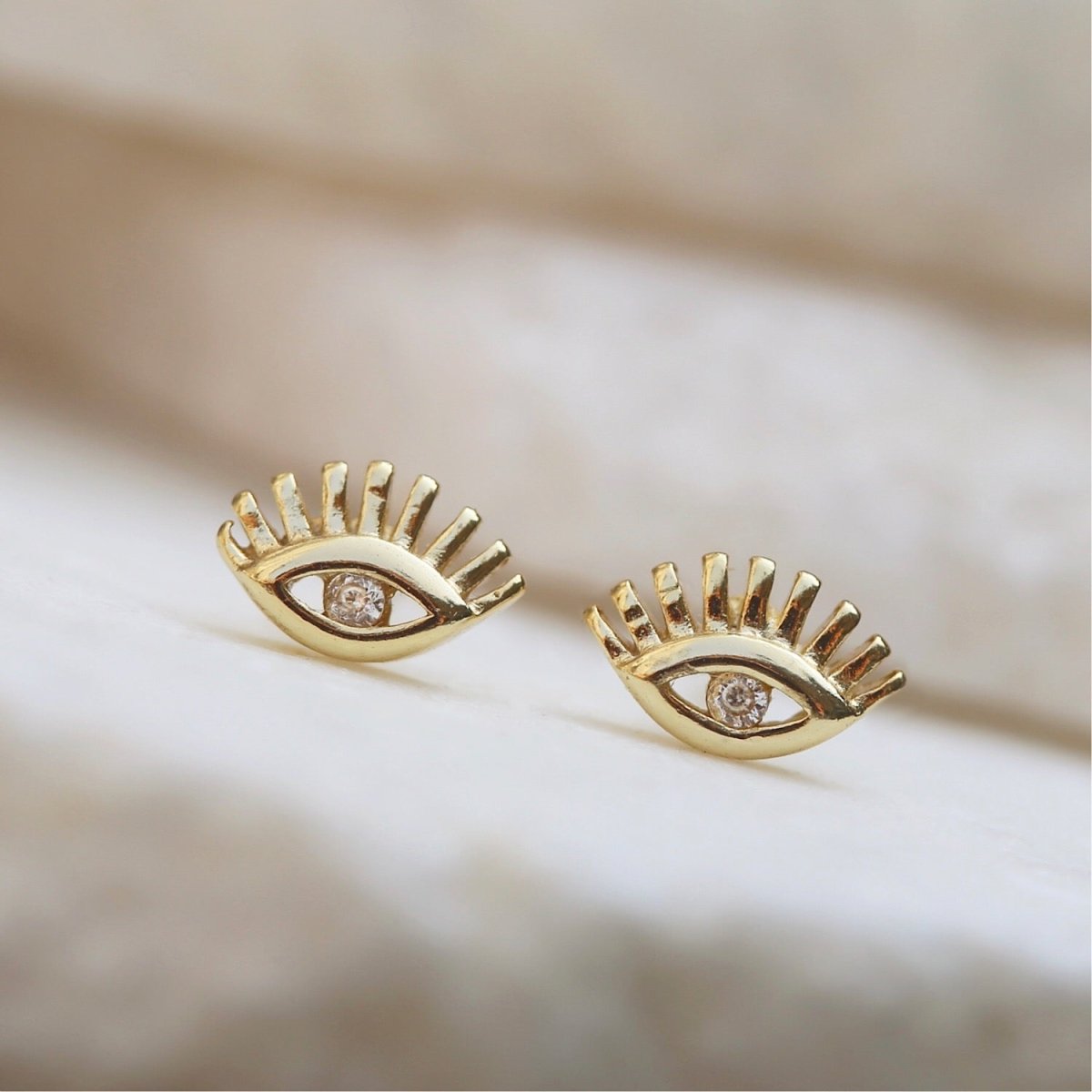 Gold vermeil evil eye studs with a clear center stone, close-up detail on a cream background