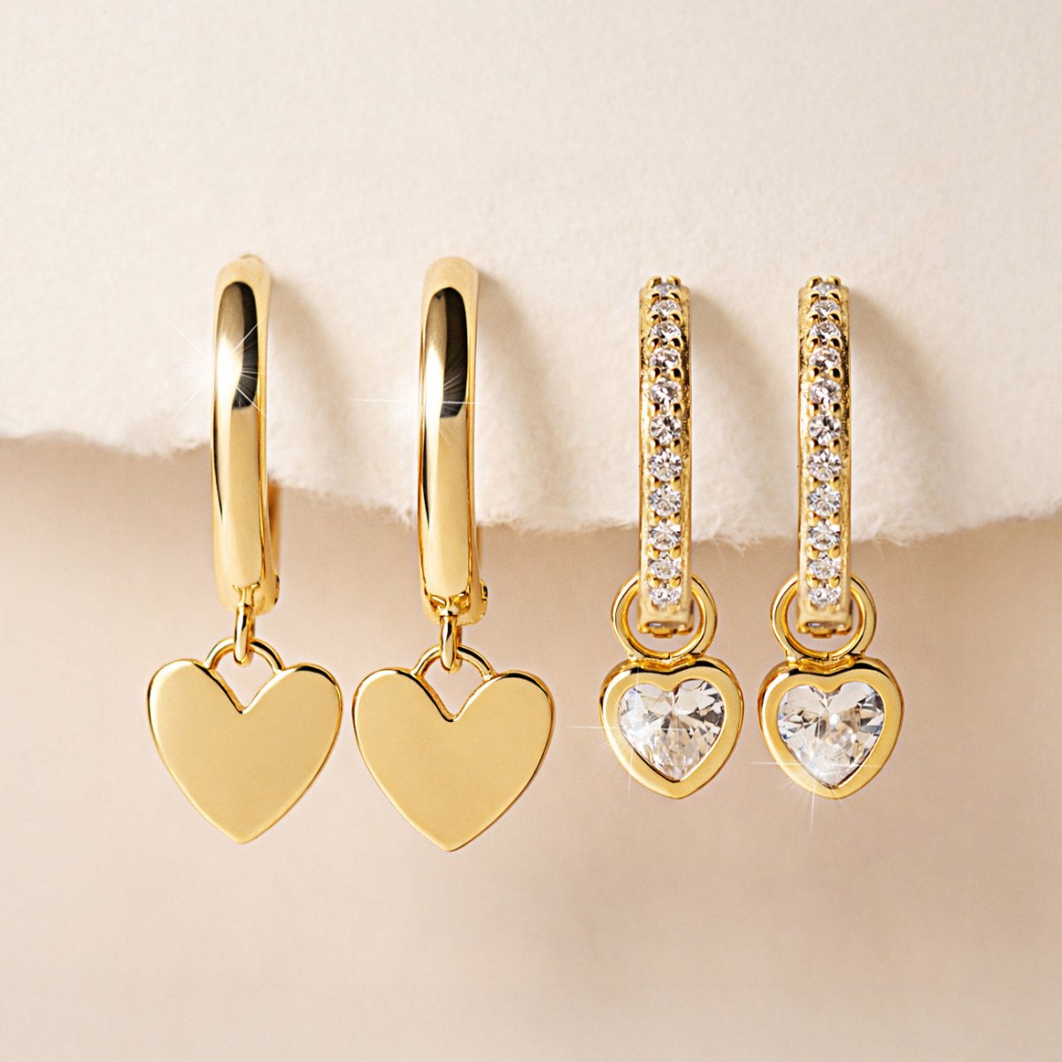 Gold vermeil huggie hoops with plain heart charms and clear-stone heart charms, on a cream background