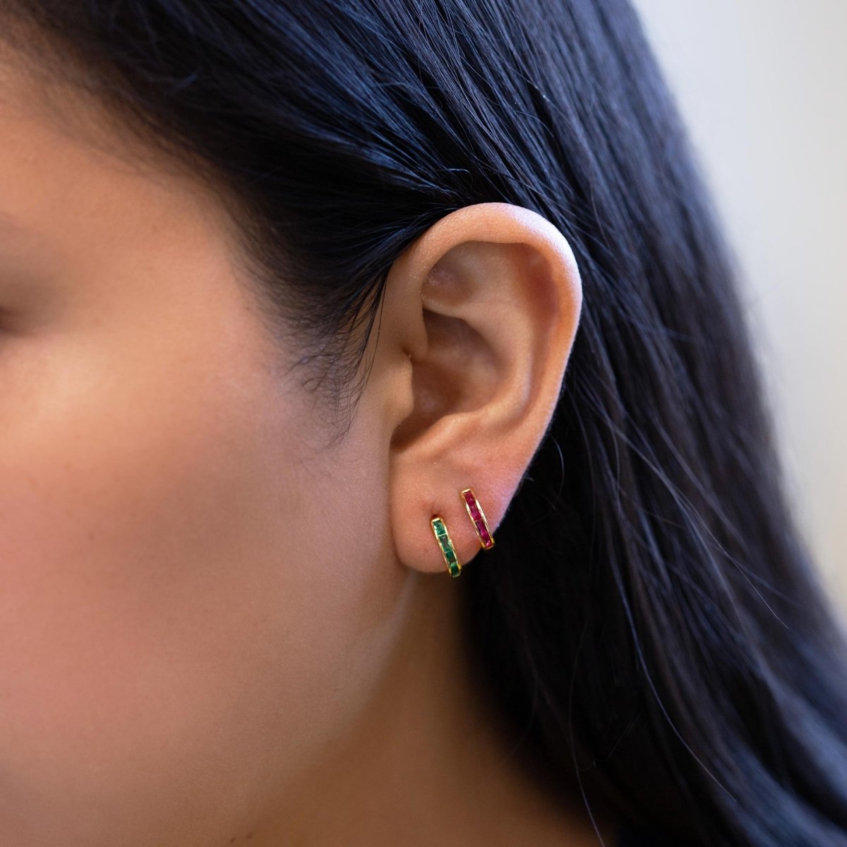 Gold vermeil ruby and green-stone baguette huggie hoops shown side by side on a model's lobe