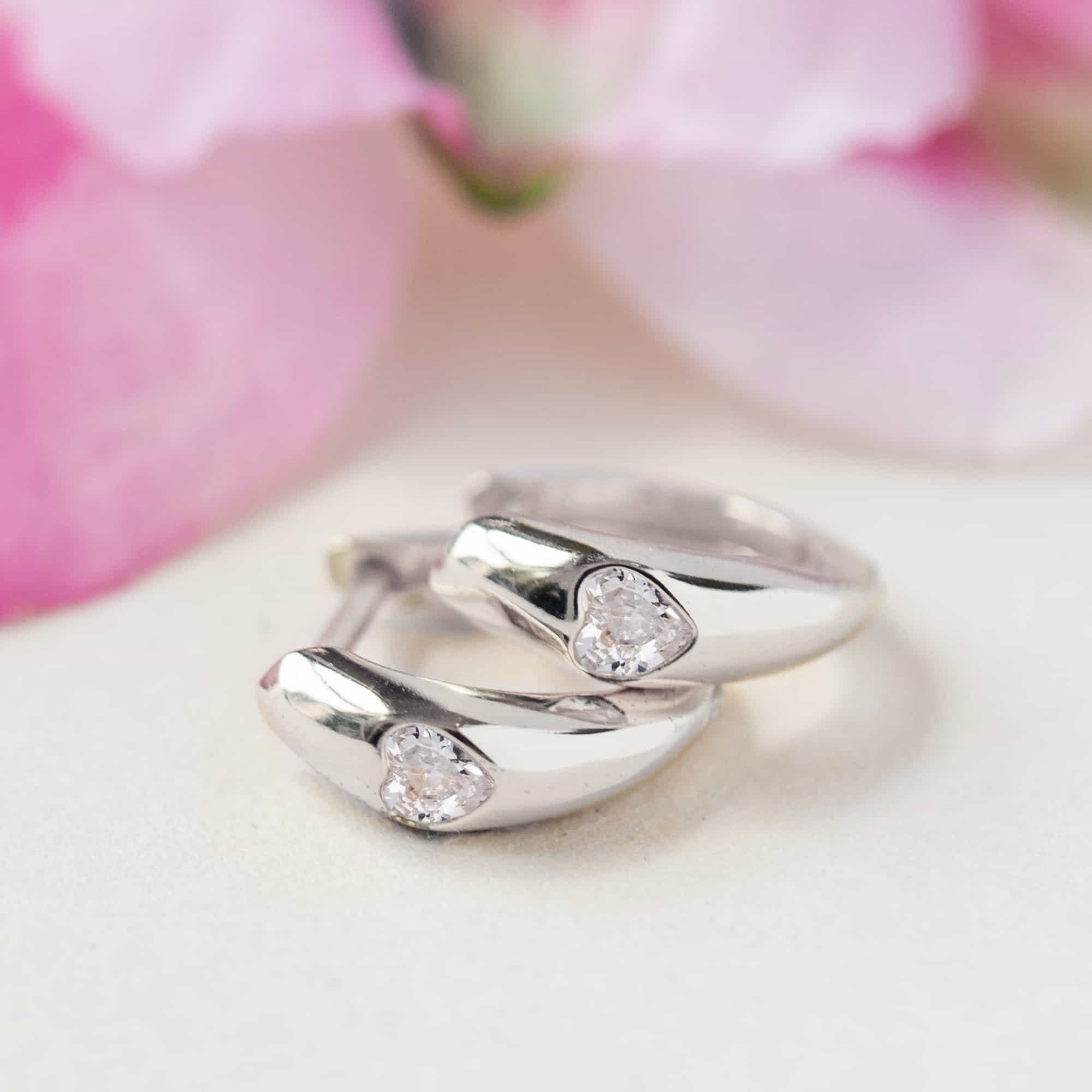 Sterling silver heart huggie hoops set with clear zirconia hearts, close-up detail on a cream background