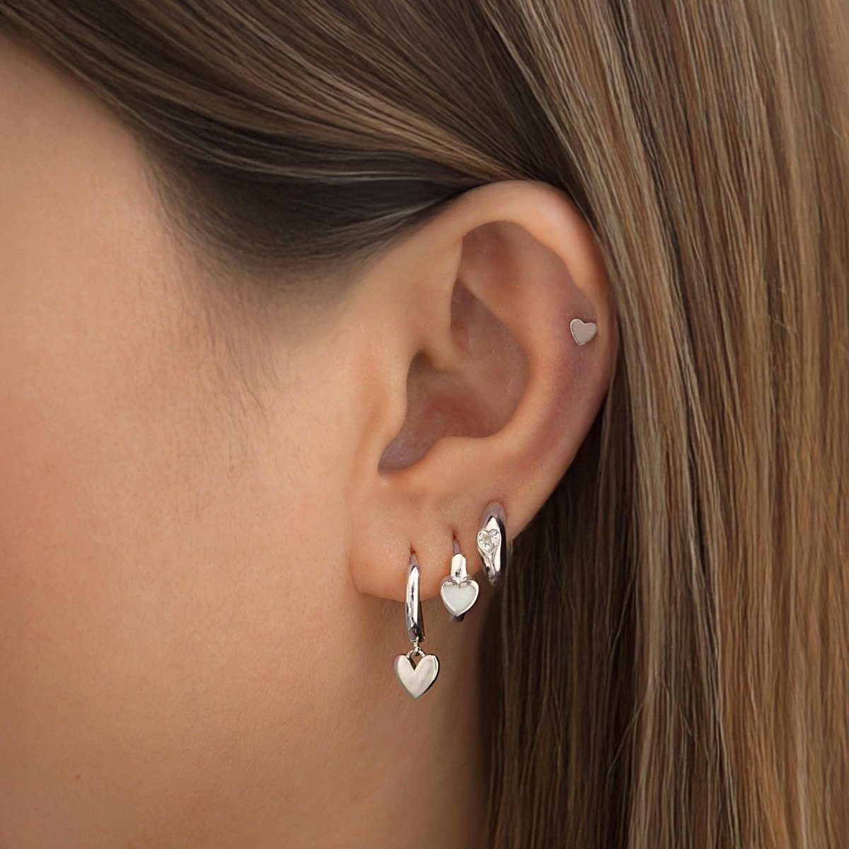 Silver heart charm huggie hoops and a tiny heart stud shown stacked on a model's ear