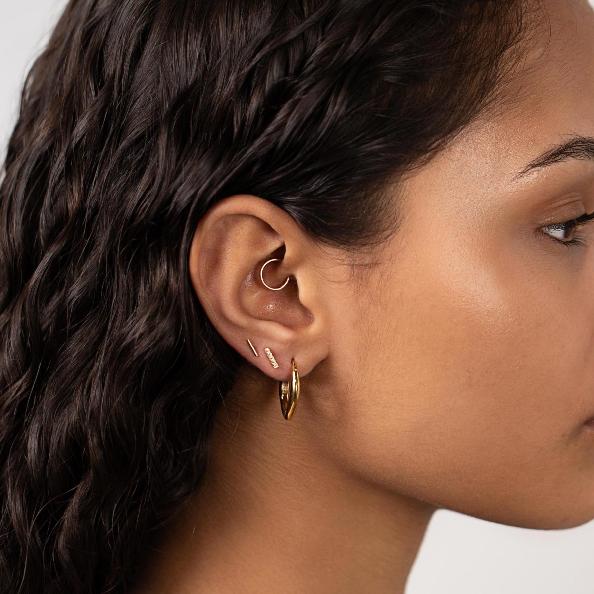 Gold vermeil thin bar stud shown on a model's lobe with hoops and a pavé bar