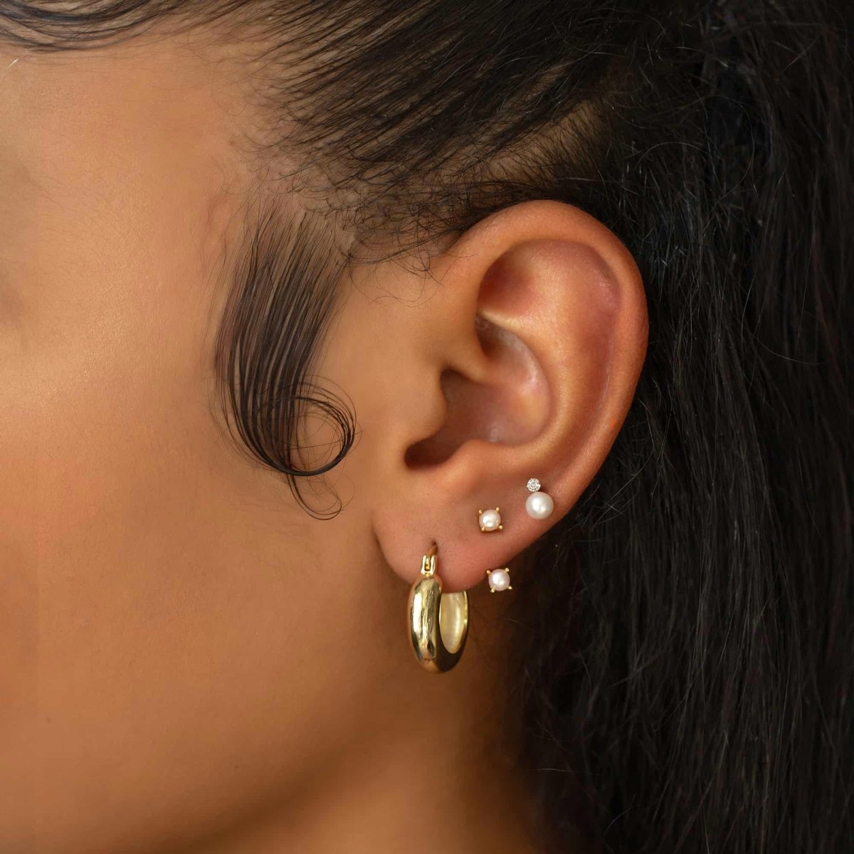 14K gold pearl stud shown on a model's lobe in a stack with a chunky gold hoop and studs