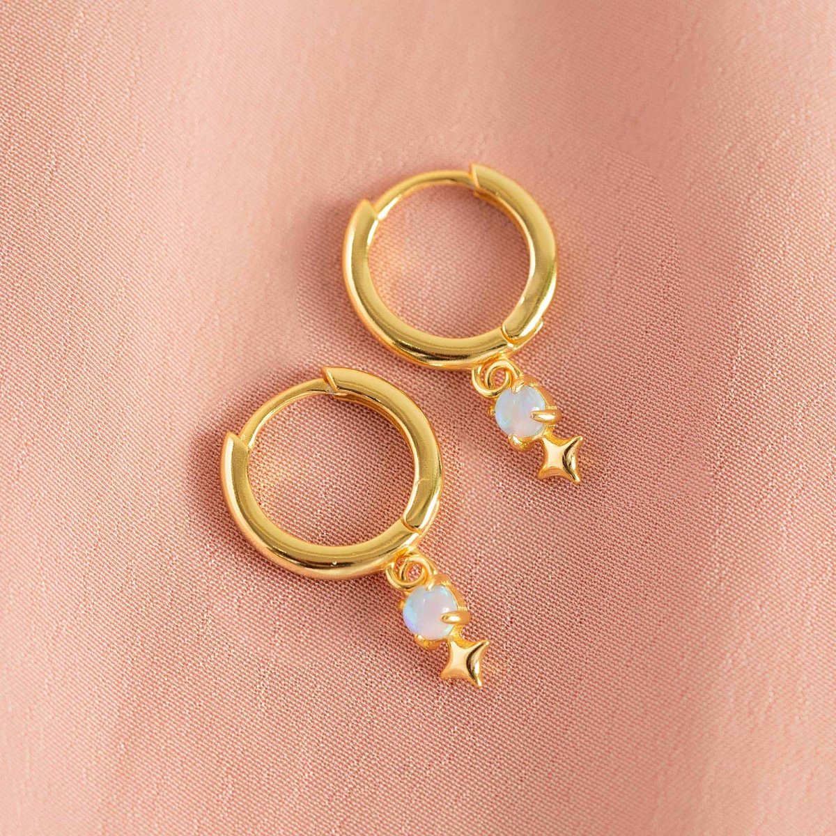 Gold vermeil huggie hoops with a light blue opal and star drop, on a pink fabric background