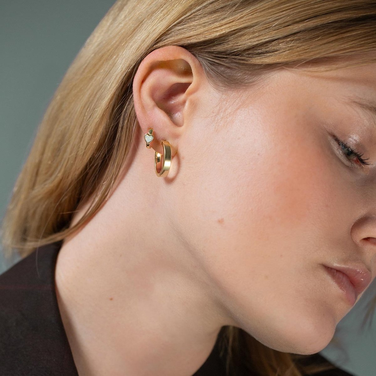 Gold wide hoop and a small heart charm huggie shown stacked on a model's lobe