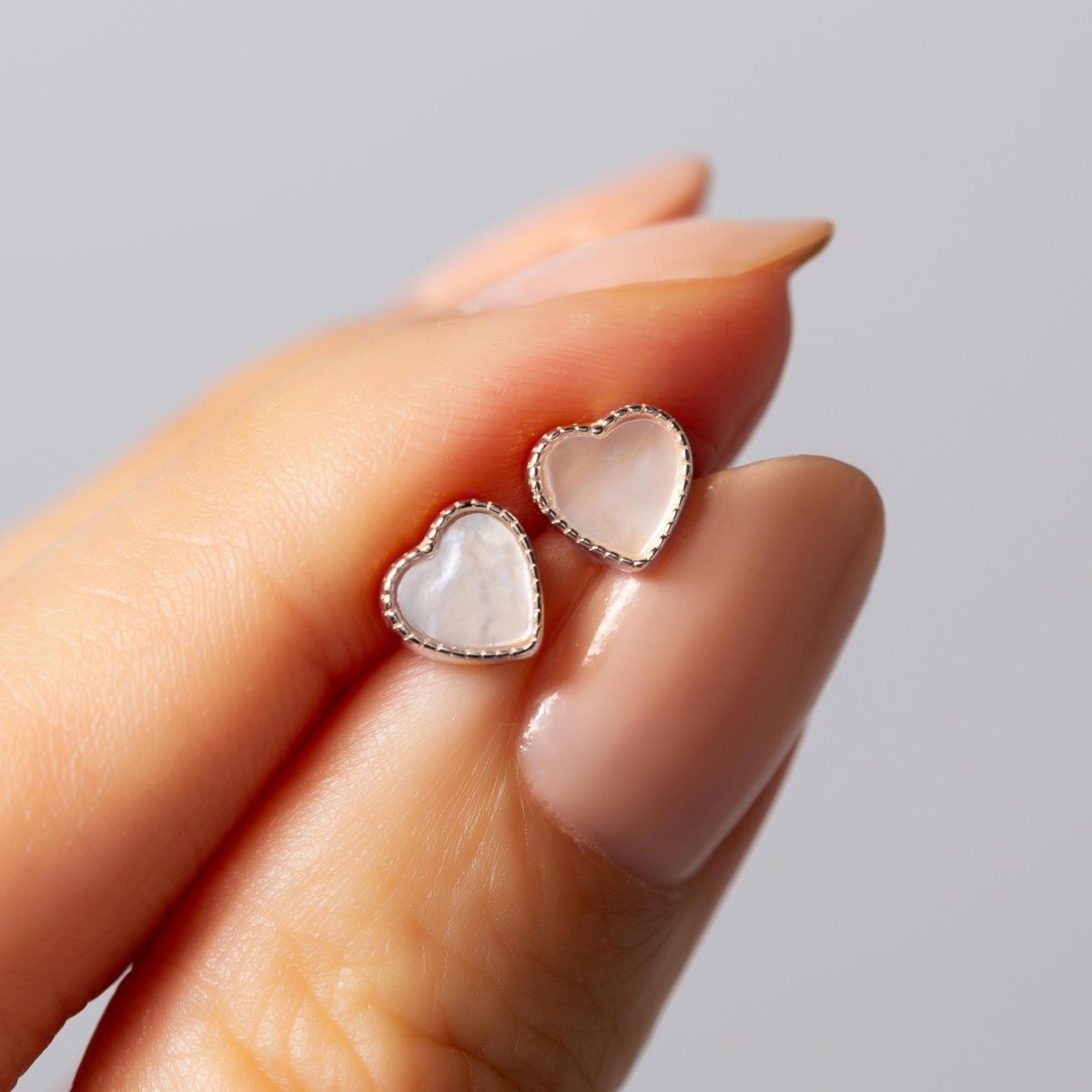 Sterling silver mini mother of pearl heart studs held between fingertips for scale