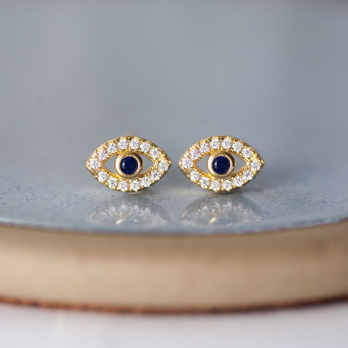 Pair of gold vermeil evil eye studs with blue sapphire centers and clear stones, on a gray dish