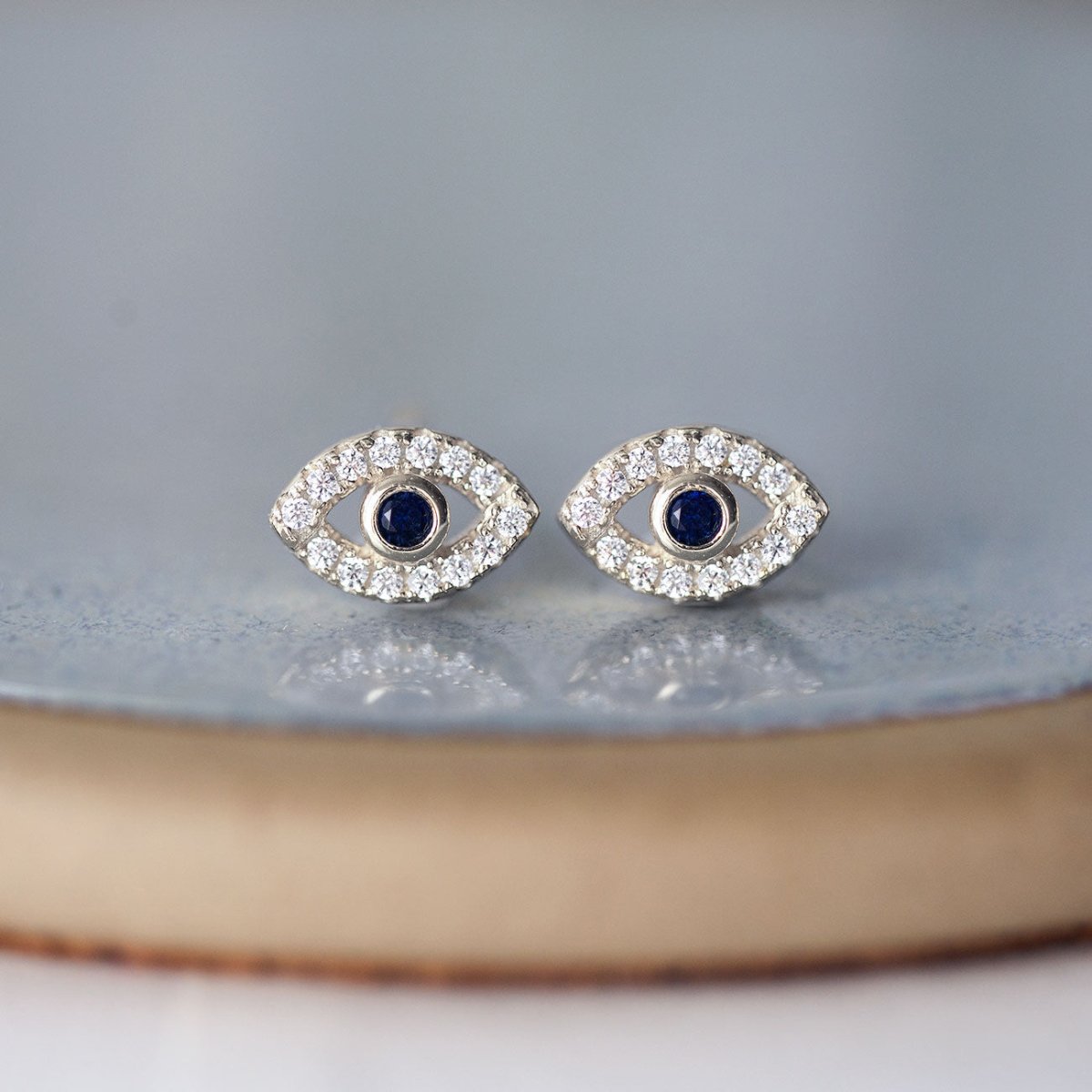 Pair of sterling silver evil eye studs with blue sapphire centers and clear stones, on a gray dish