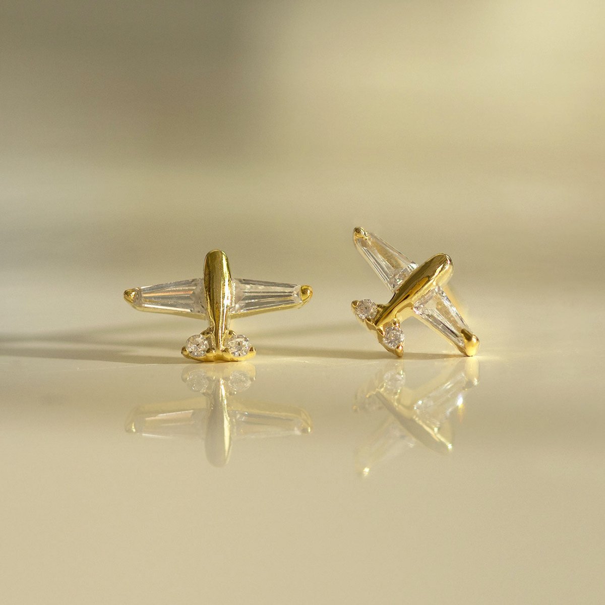 Gold vermeil airplane studs with clear baguette wings, shown at two angles with a soft reflection
