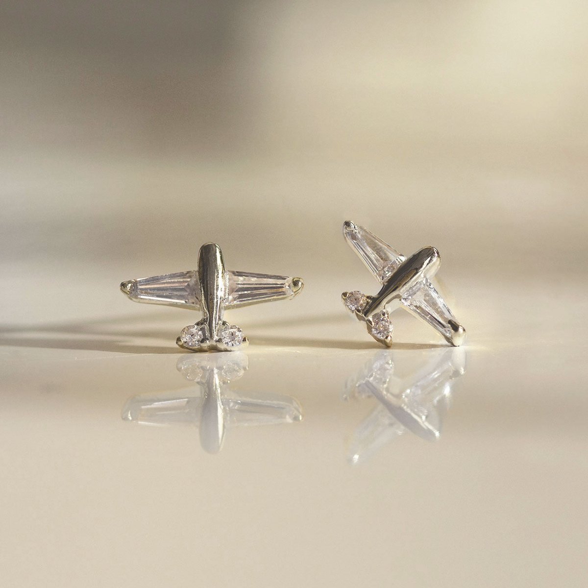 Sterling silver airplane studs with clear baguette wings, two angles on a warm reflective surface