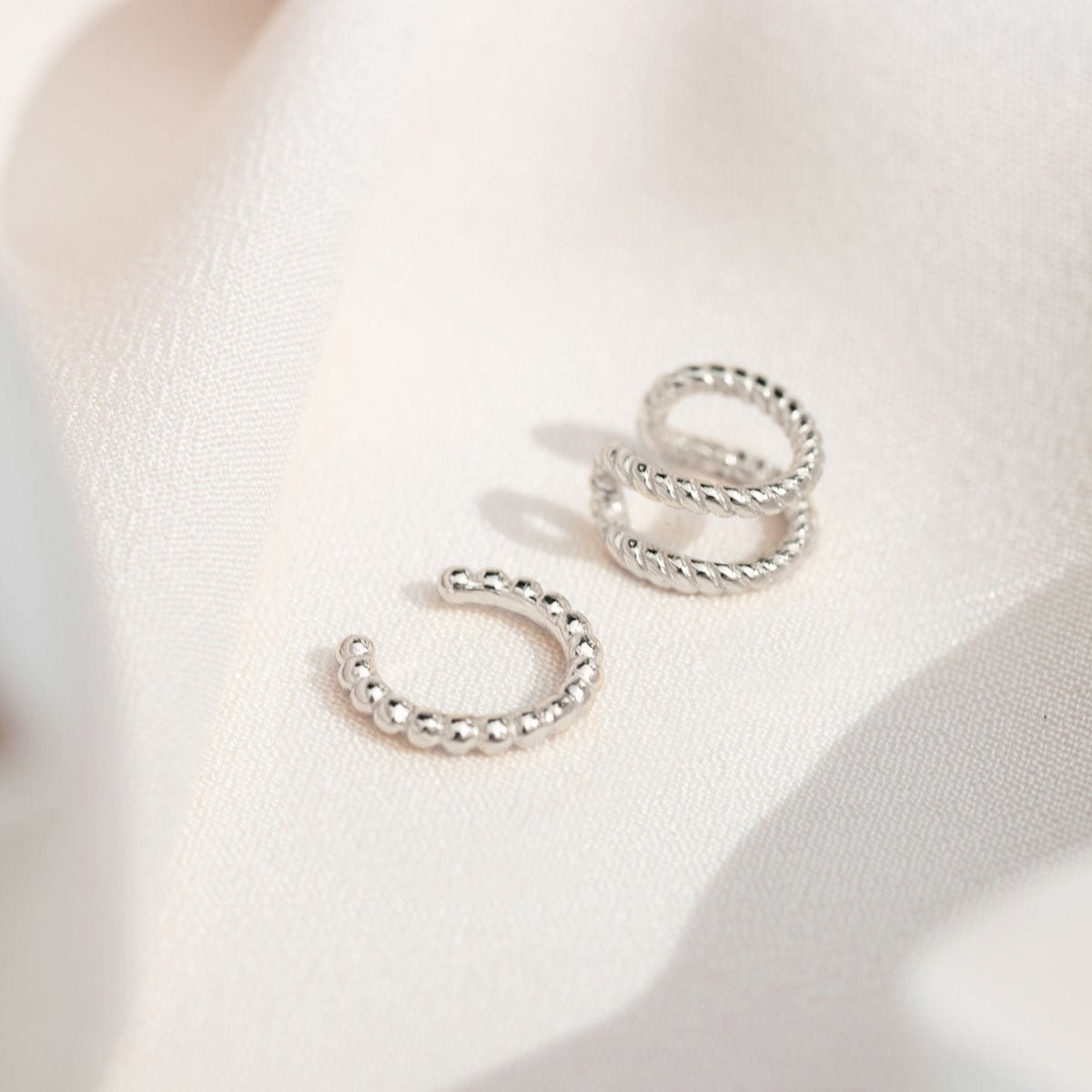 Sterling silver beaded ear cuff beside a twisted double-band cuff, laid on cream fabric